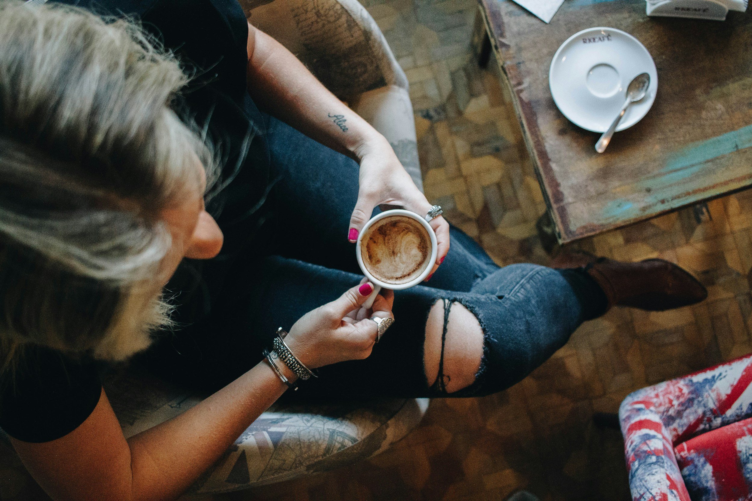 A woman with blonde hair, wearing multiple bracelets and a black shirt, sitting on a patterned armchair, holding a cup of coffee, with a torn black tank top, ripped jeans, and brown boots. There is a small wooden table with an empty cup and spoon on it next to her.