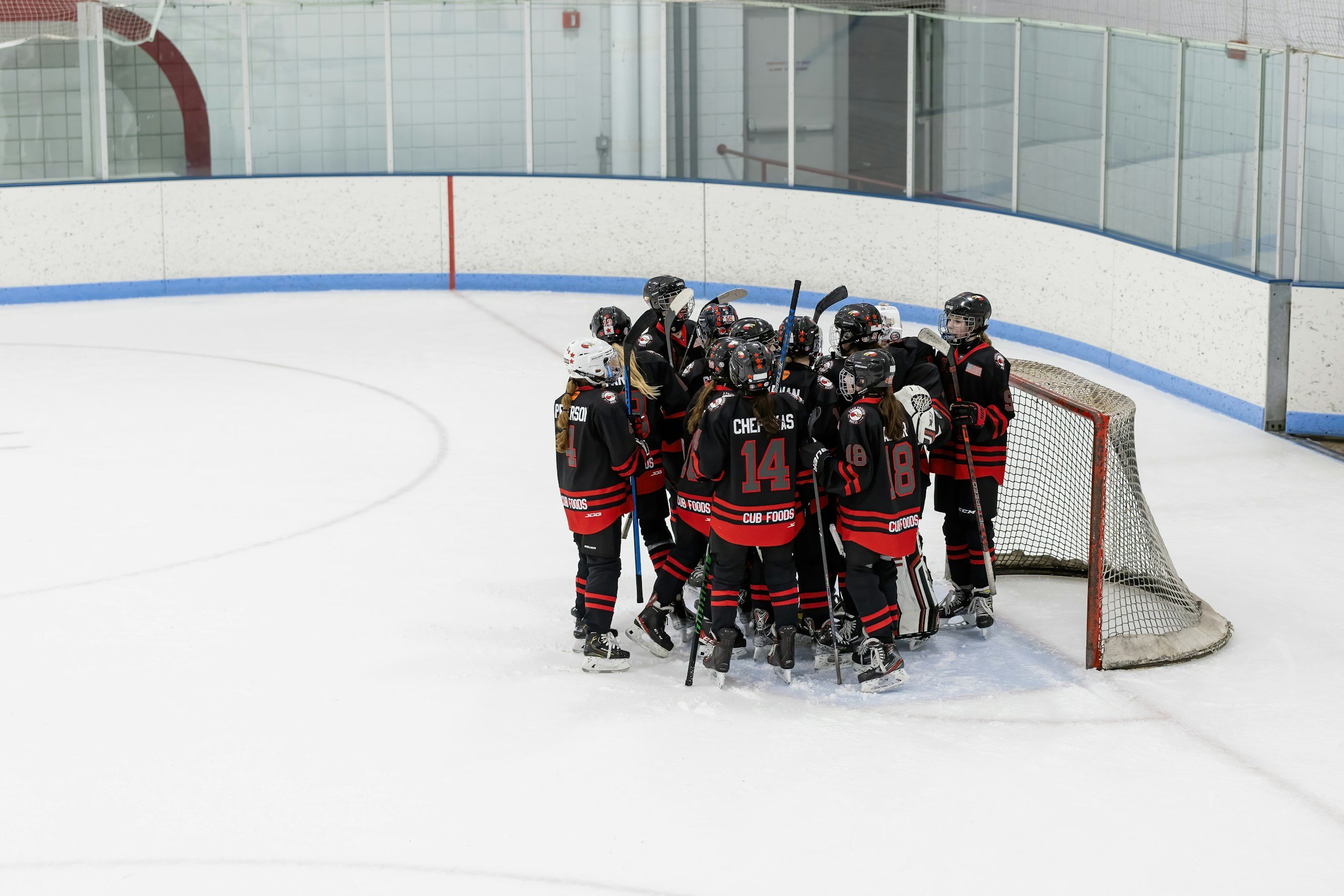 A group of young ice hockey players gathered together near the goalpost on the ice rink, celebrating or discussing during a game or practice. They are dressed in black and red uniforms with helmets and hockey sticks.