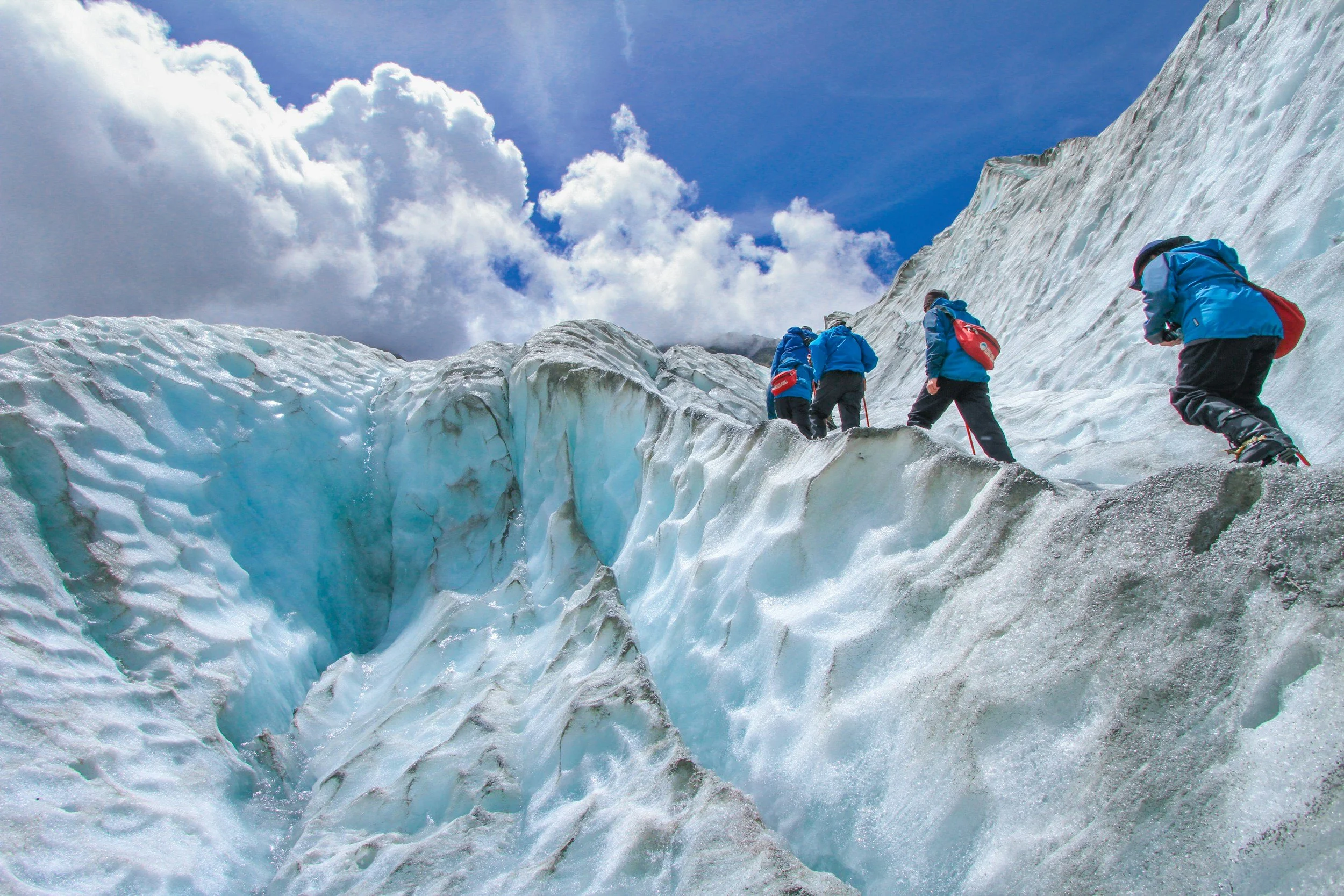 Four climbers in blue jackets ascending a large icy glacier under a partly cloudy sky.