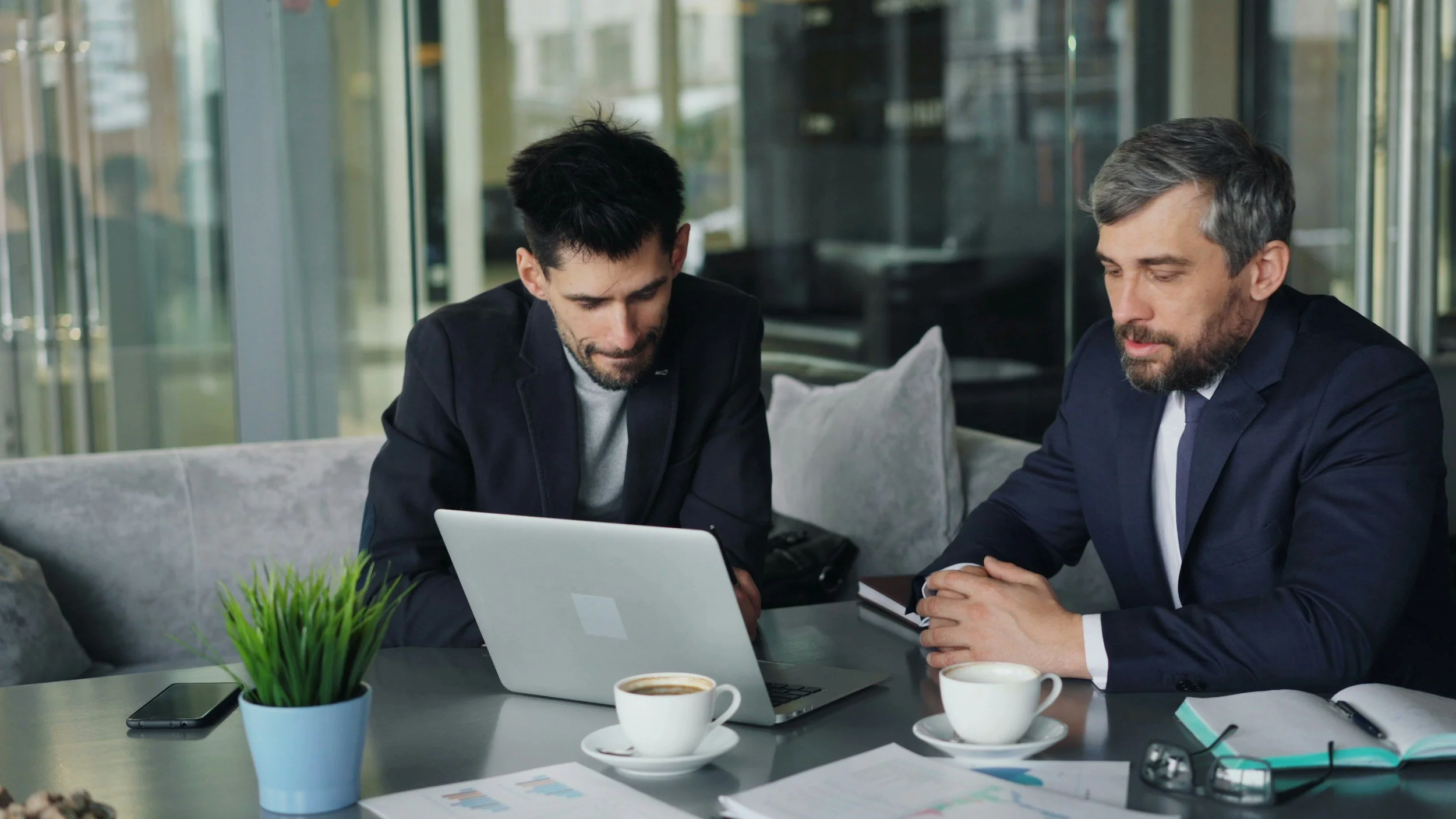 Two men in business attire sitting at a table in a modern office, looking at a laptop screen. The table has coffee cups, a potted plant, and documents.