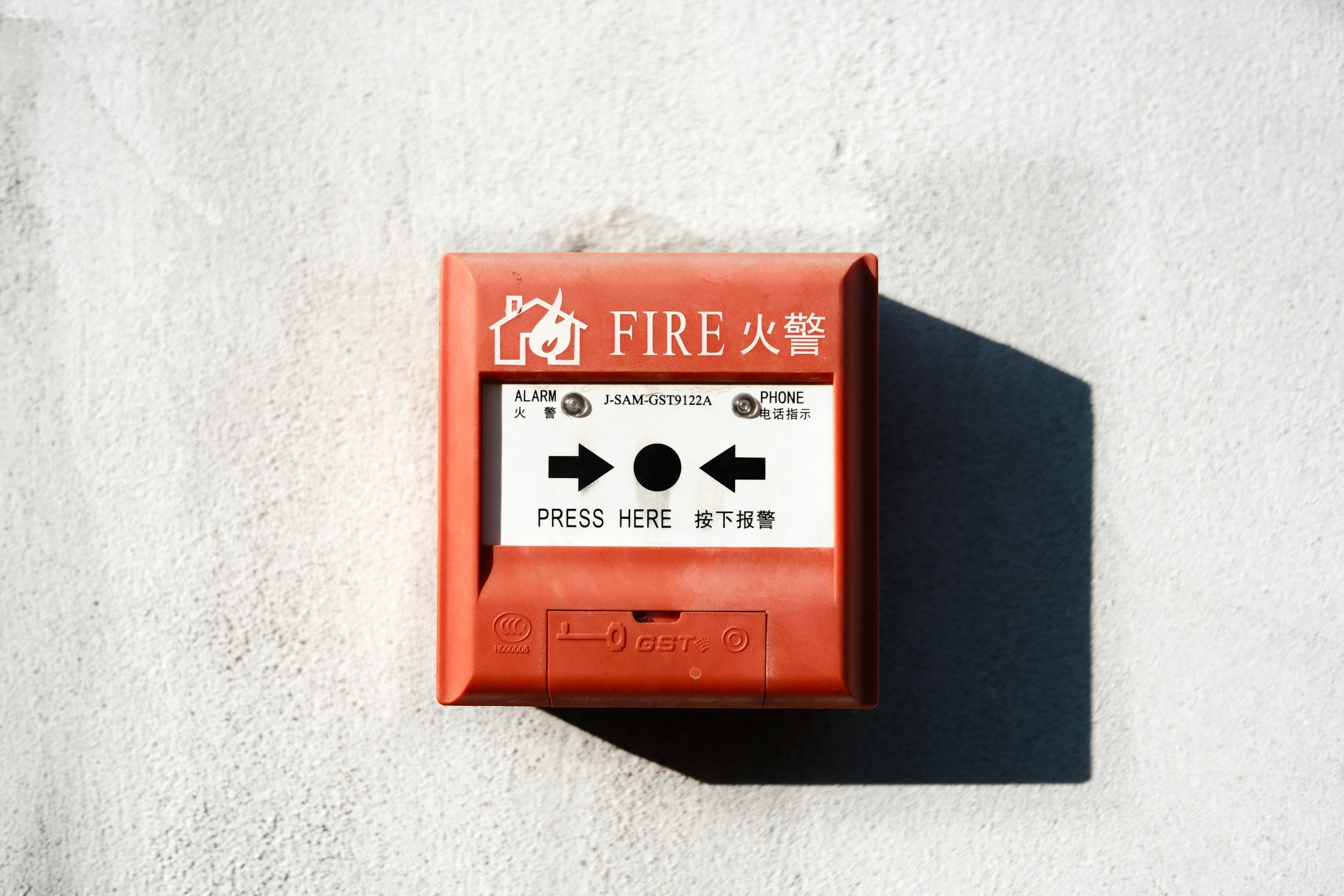 Red fire alarm box mounted on a white wall with instructions in English and Chinese, and directional arrows pointing to a button labeled "PRESS HERE."