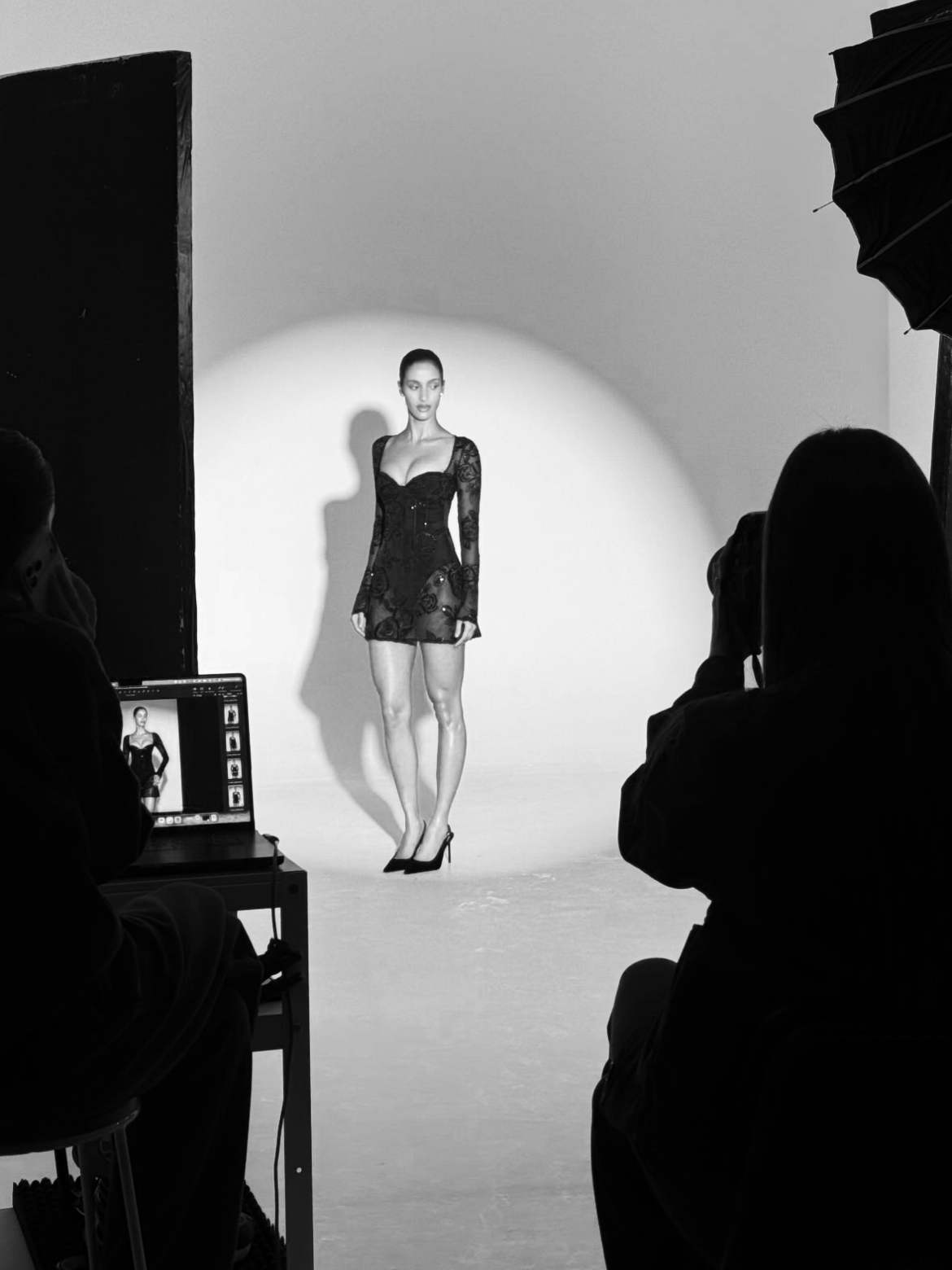 A black-and-white photo of a fashion photoshoot studio. A model wearing a black lace dress and high heels stands in front of a plain white background, with photographers and crew members around her. behind the scenes