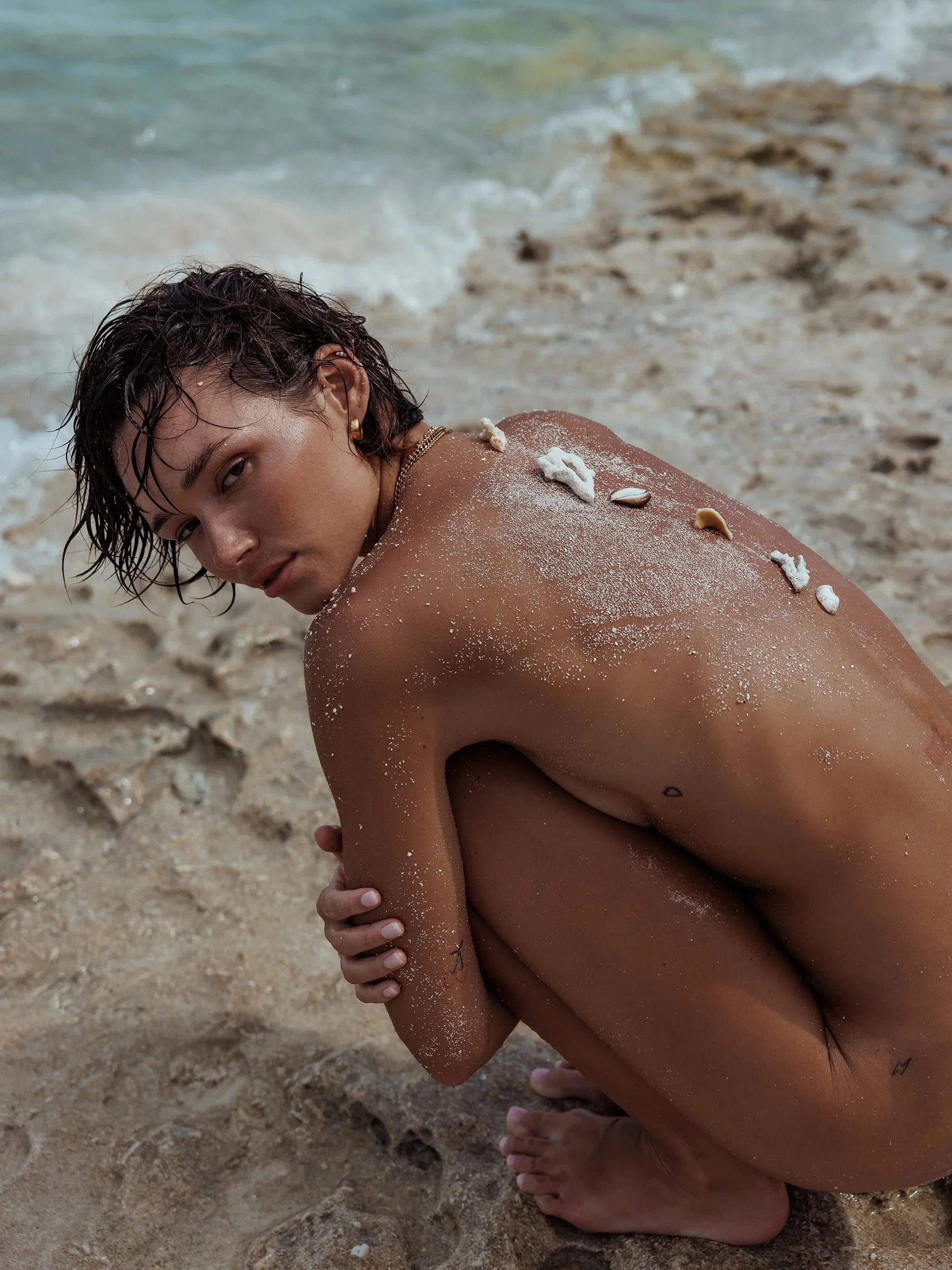 A woman with short dark wet hair squats on a sandy beach near the ocean, with seashells and white sand on her back, looking over her shoulder at the camera.