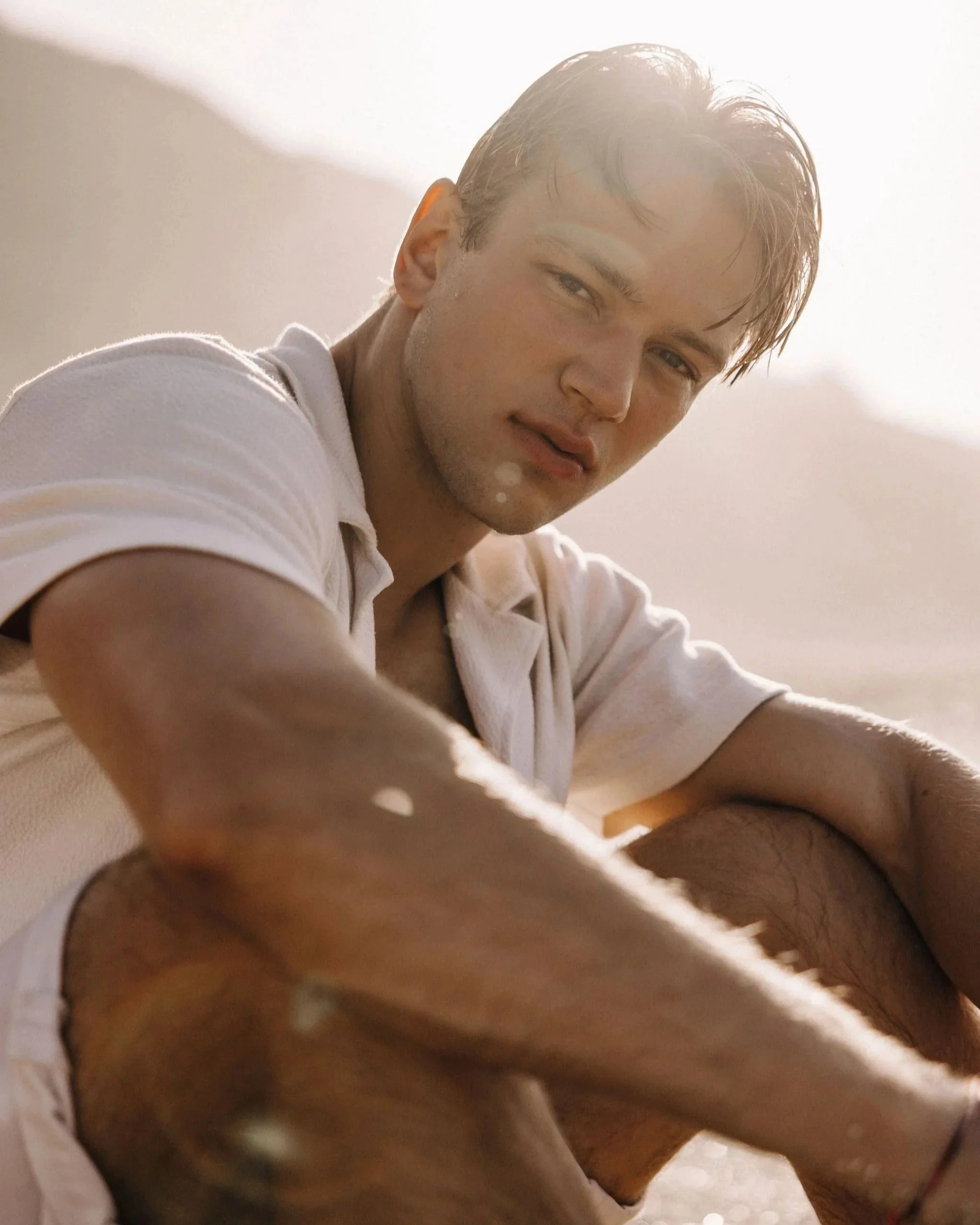 A young man sitting outdoors in sunlight, wearing a white shirt and looking at the camera.