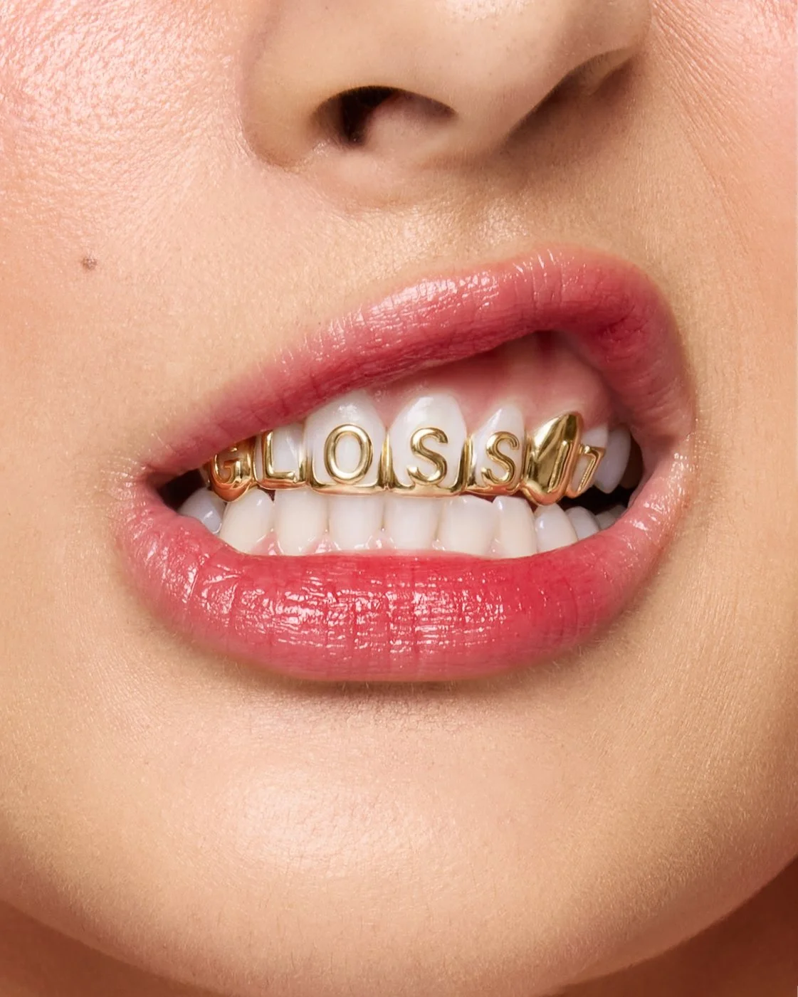 Close-up of a woman's mouth with pink lipstick, showing white teeth with a gold dental grill that spells 'GLOSSY' in capital letters.