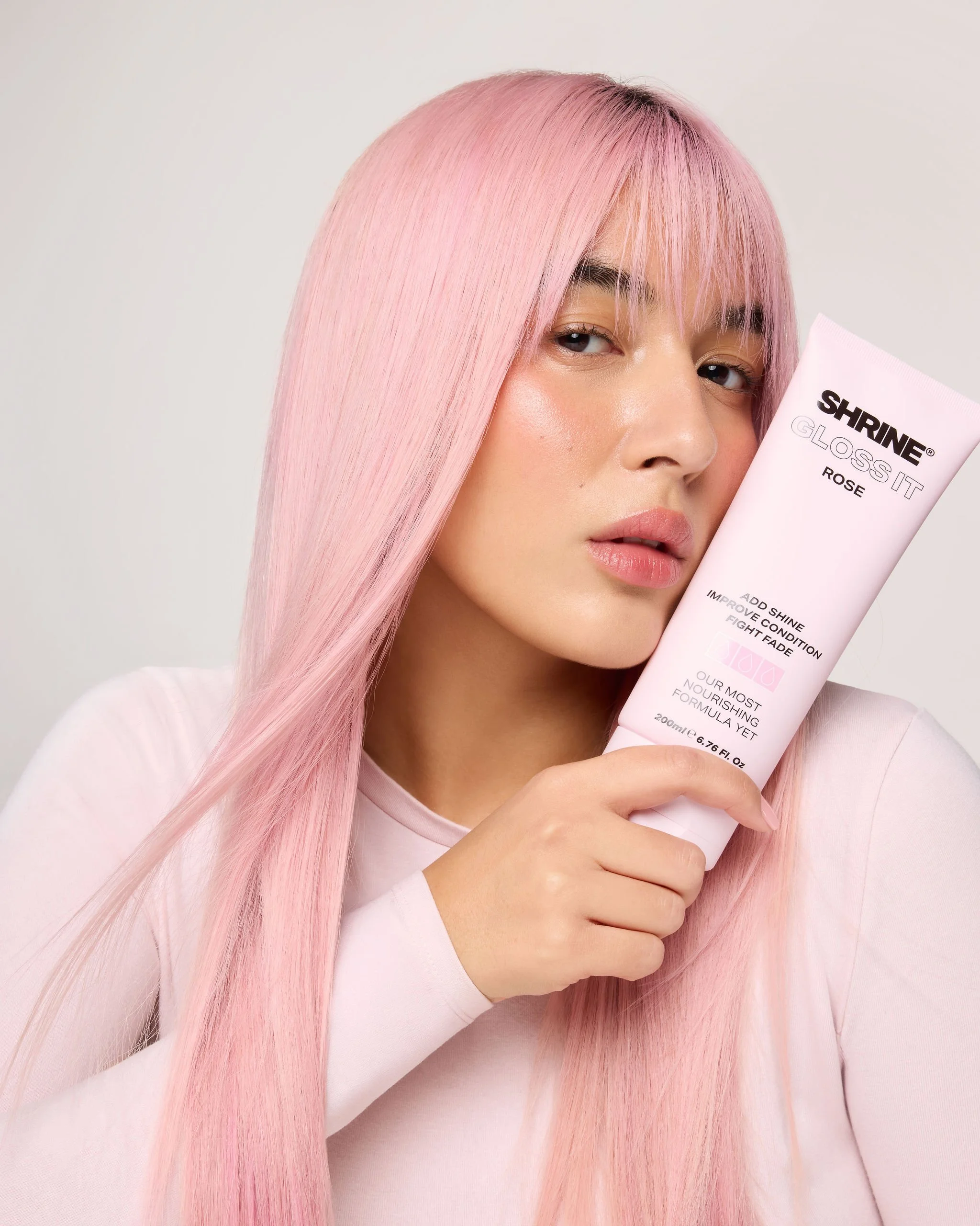 A woman with long, straight pink hair holding a tube of Shrine Gloss It in Rose color close to her face, with a neutral background.