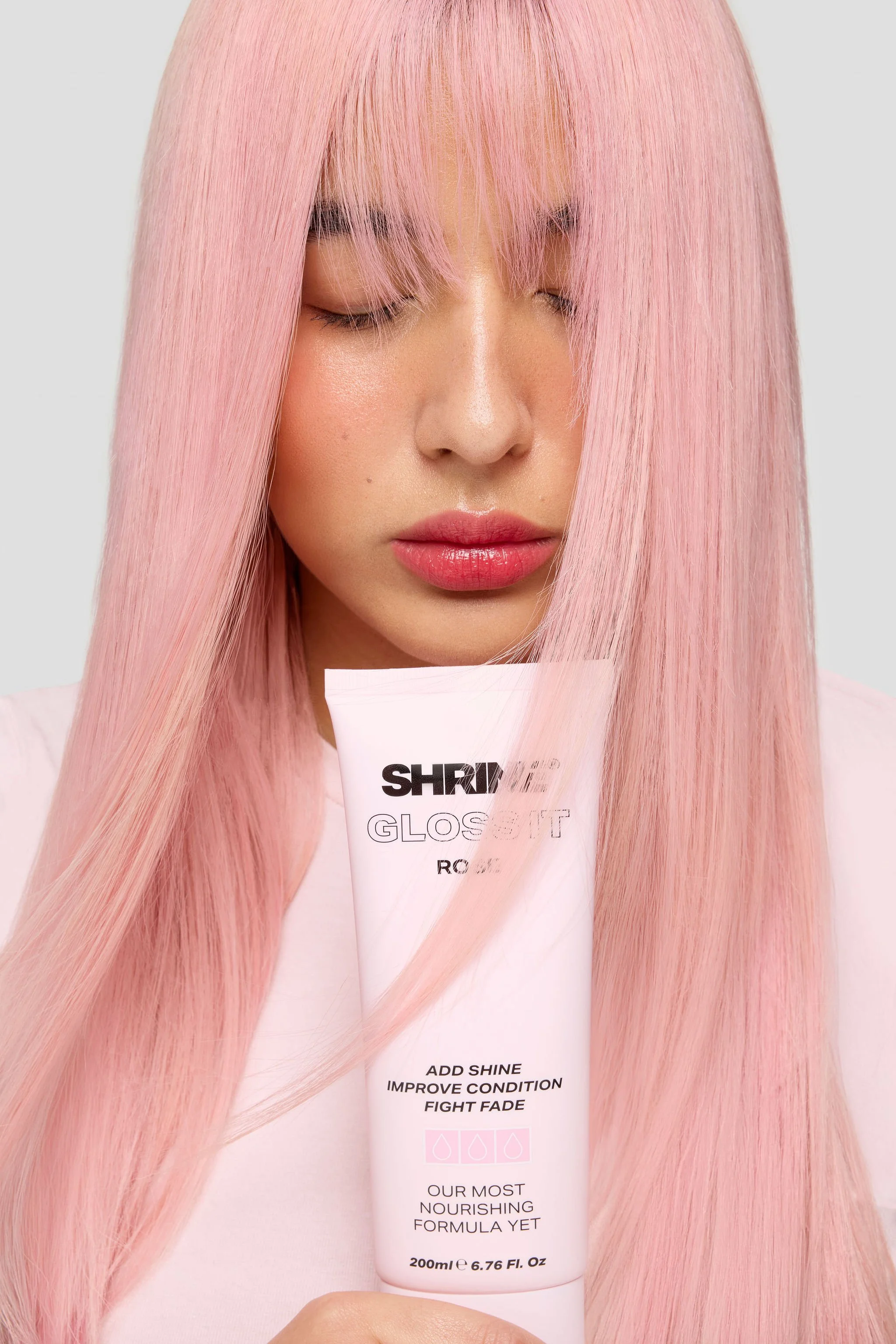 A woman with long, straight pink hair, closed eyes, and pink lipstick holding a tube of Shrink Glossit product.