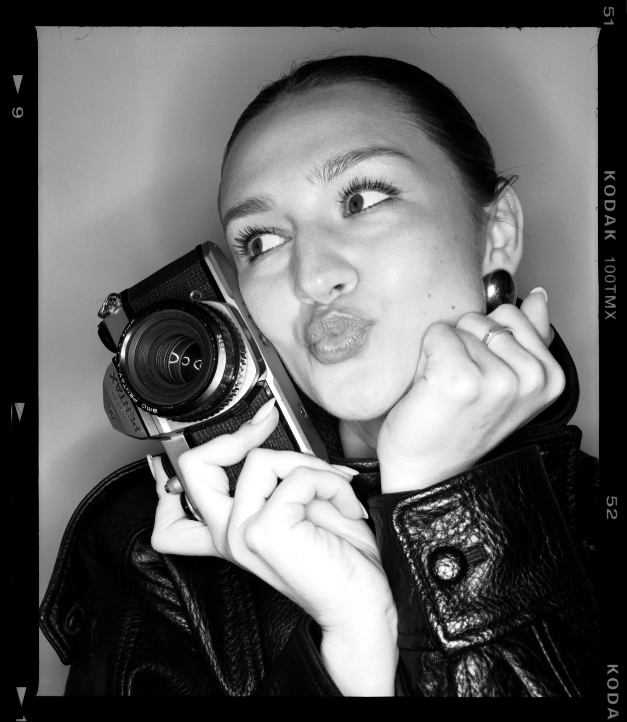 A woman with a leather jacket holding a vintage camera close to her face, making a scrunched up face and puckering her lips.