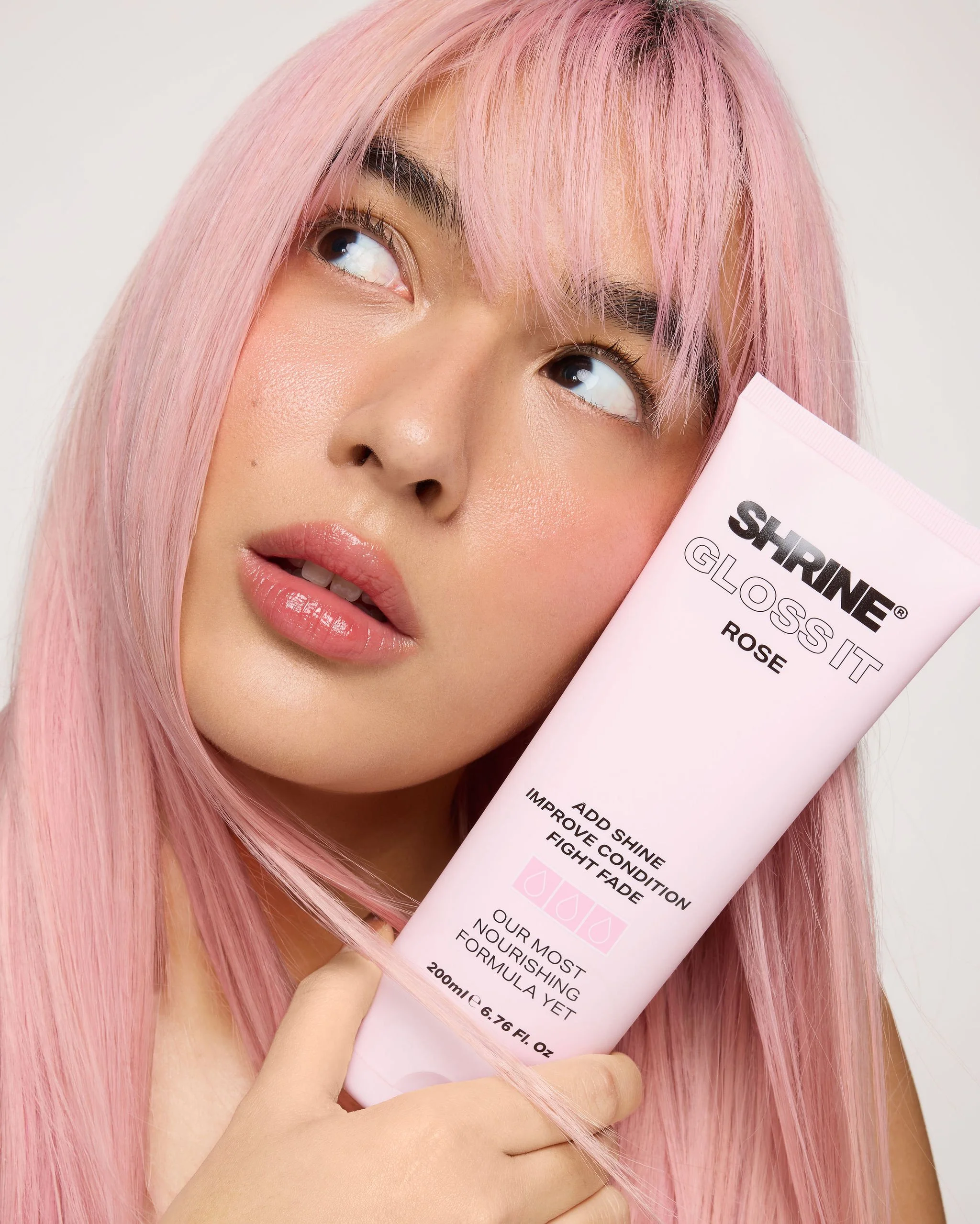 A woman with pink hair holding a tube of skincare product labeled 'Shrine Gloss It Rose' near her face.