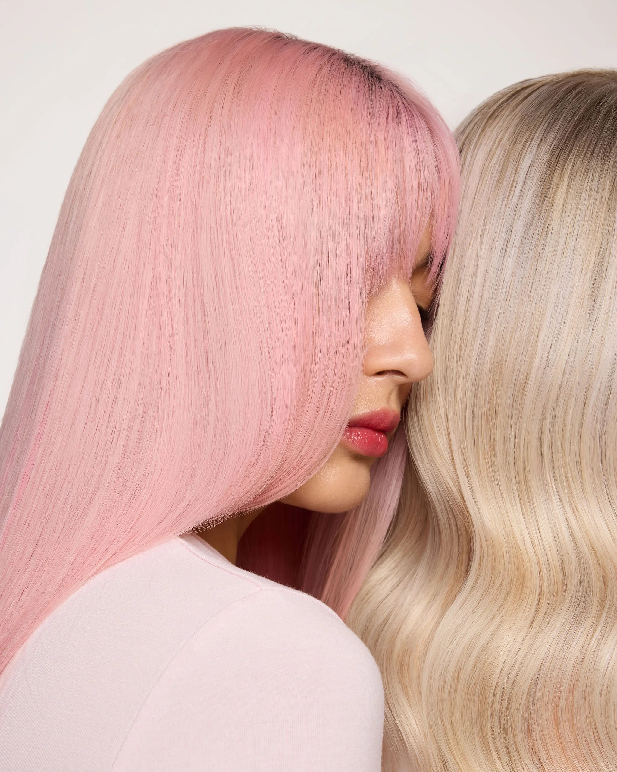 Close-up of two women with long, straight, pastel-colored hair in pink and blonde shades, side profiles, against a plain background.