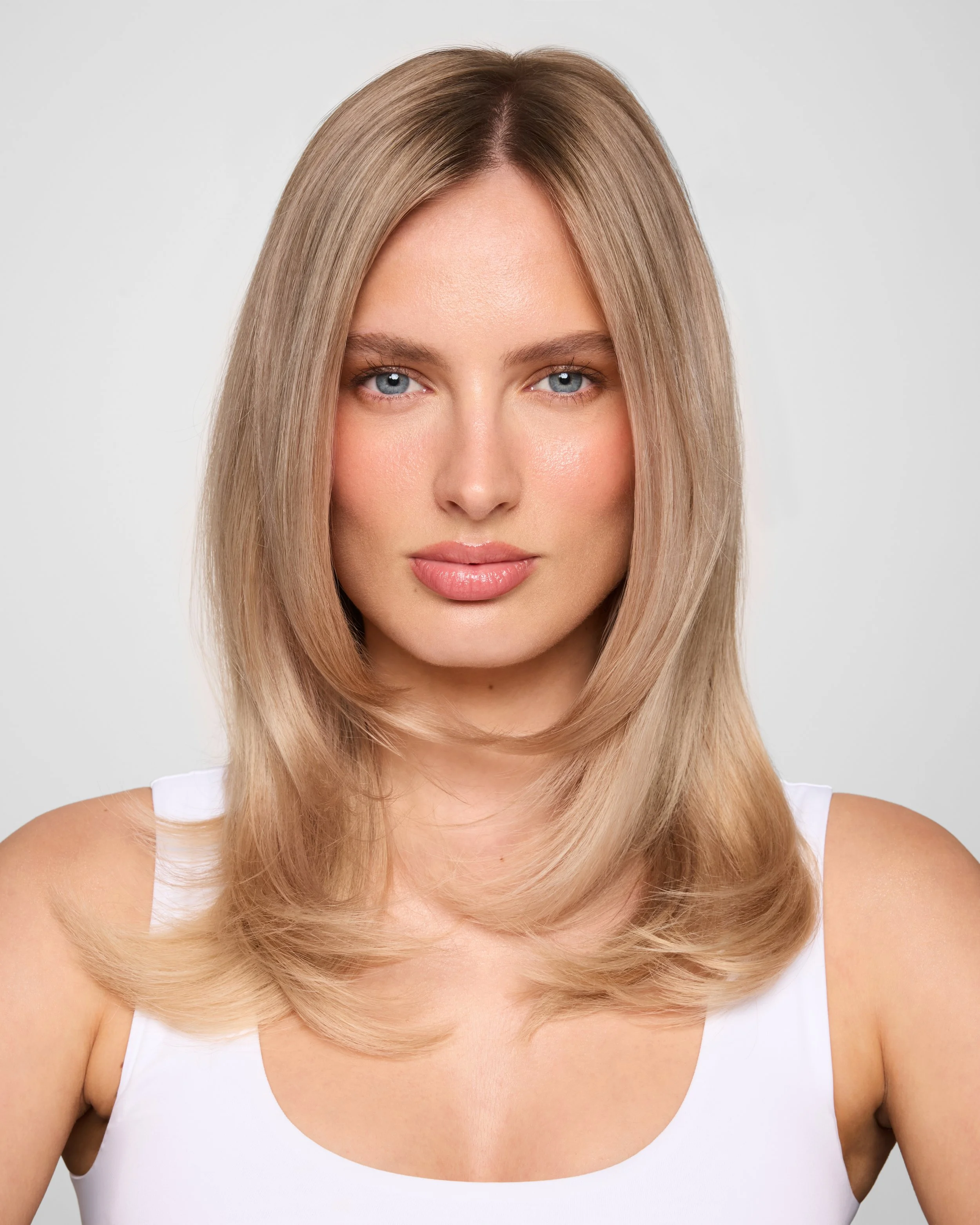 close-up beauty portrait of a blonde model with blue eyes and fair skin in a white sleeveless top against a light grey background