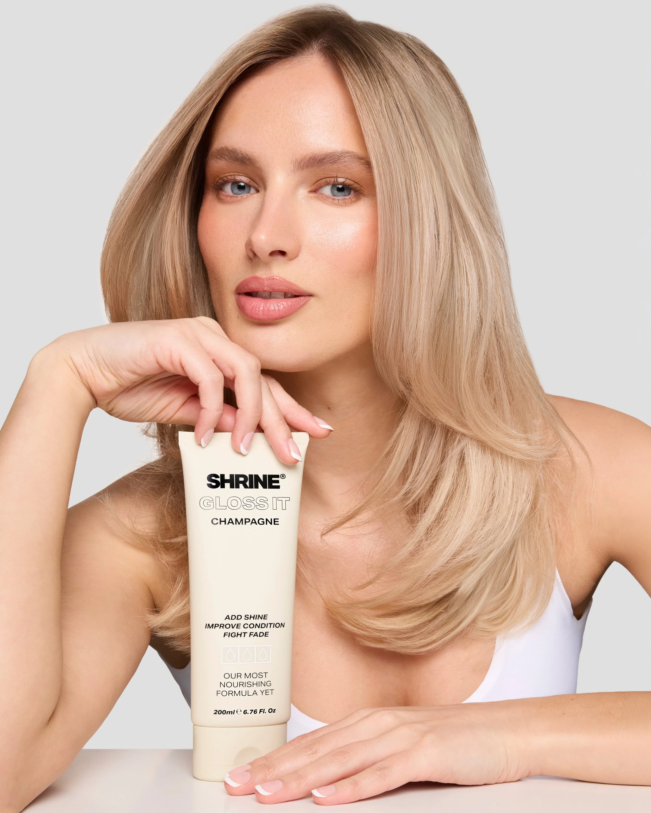 Beauty campaign shot of a model with long blonde hair and blue eyes holding Shrine Gloss It Champagne hair gloss