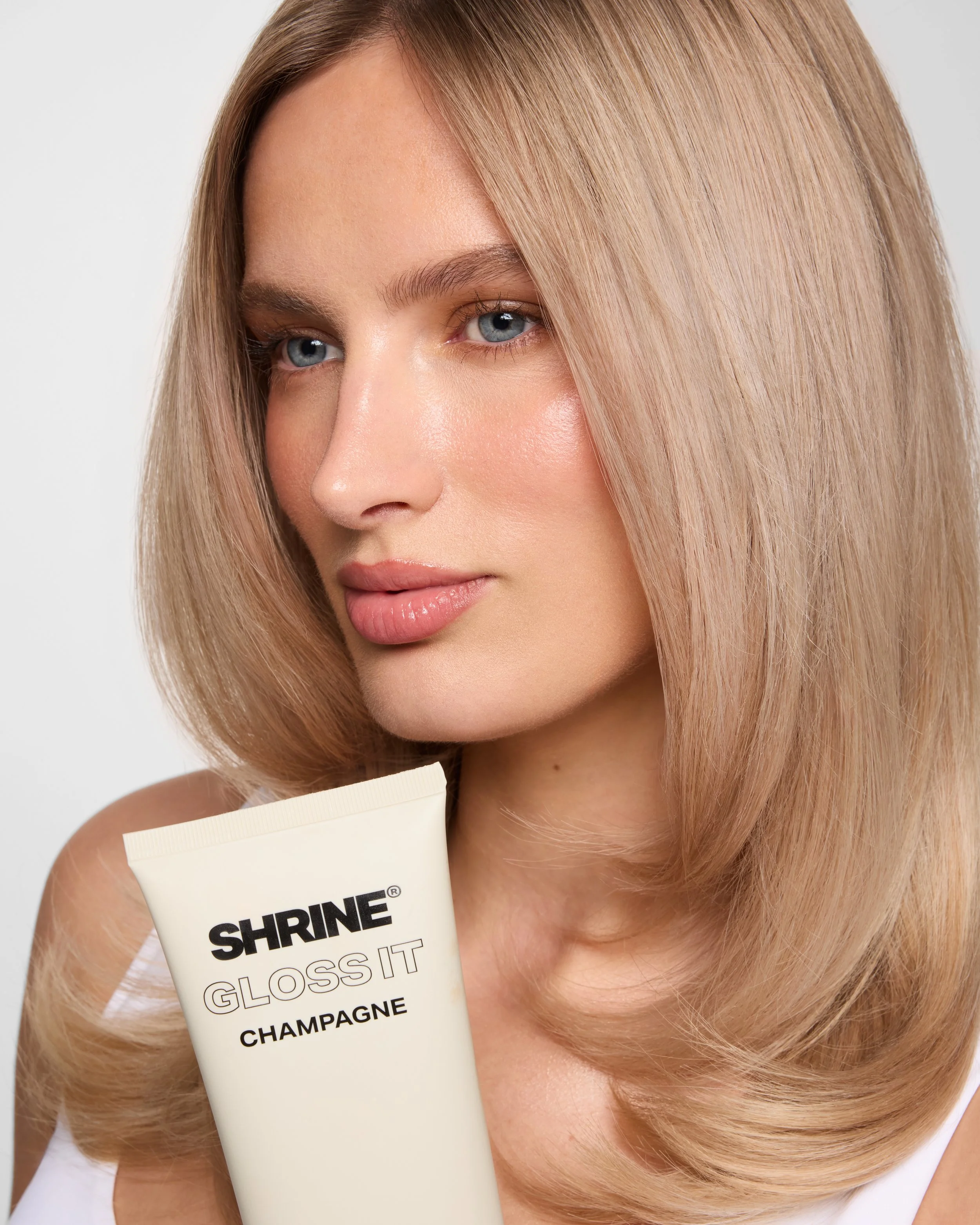 Beauty editorial close-up of a blonde model holding Shrine Gloss It Champagne product