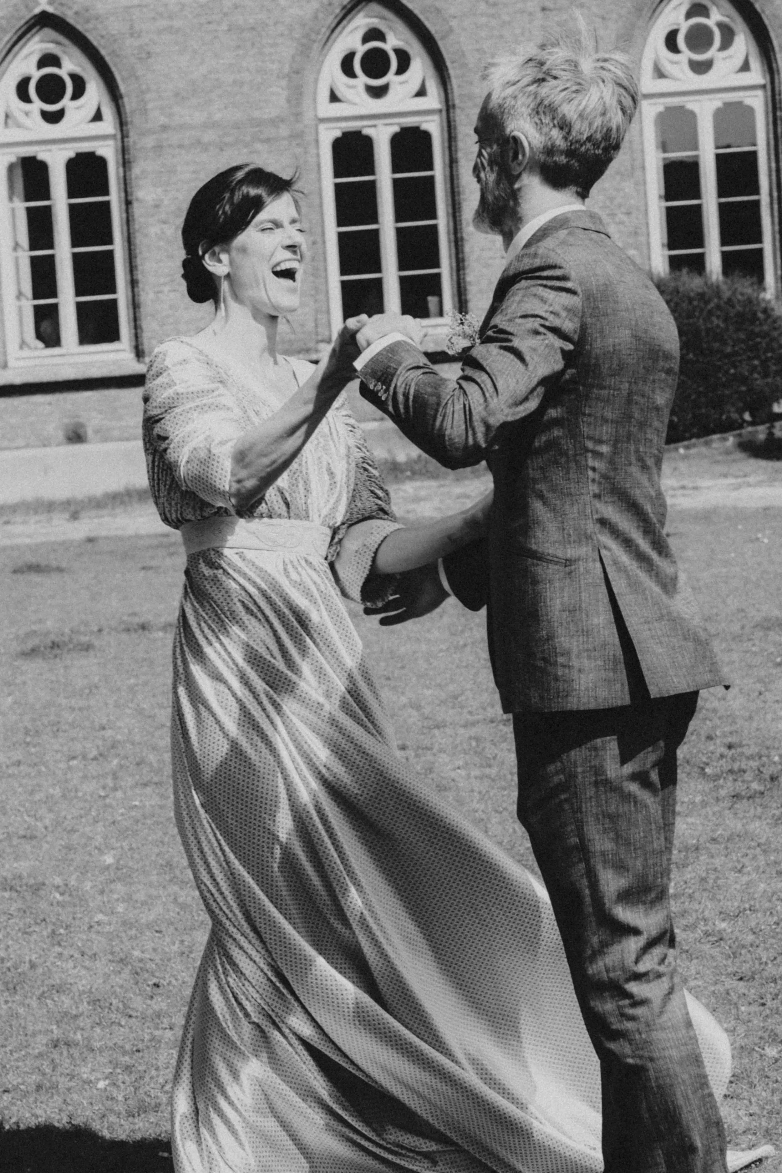 A black and white photo of a woman and a man dancing outdoors in front of a building with arched windows; the woman is smiling and wearing a vintage dress, the man is dressed in a suit.