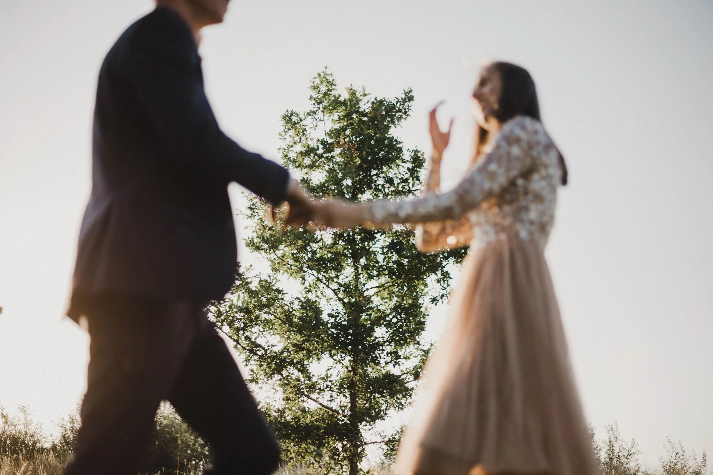 Artistieke trouwfoto van een koppel dat hand in hand danst in de velden tijdens golden hour; zij zijn zacht wazig, de achtergrond scherp