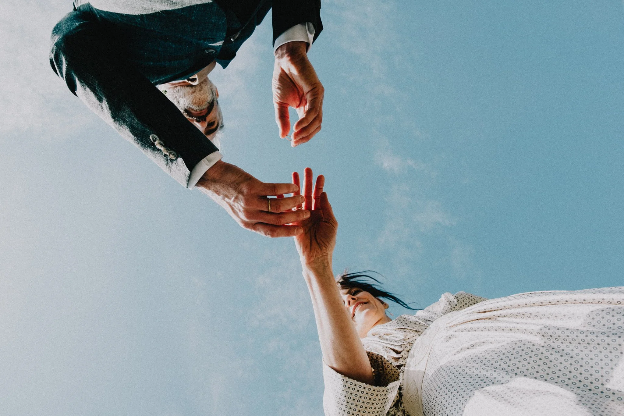 A man in a suit reaching down to shake hands with a woman in a dress against a blue sky, taken from a low angle.