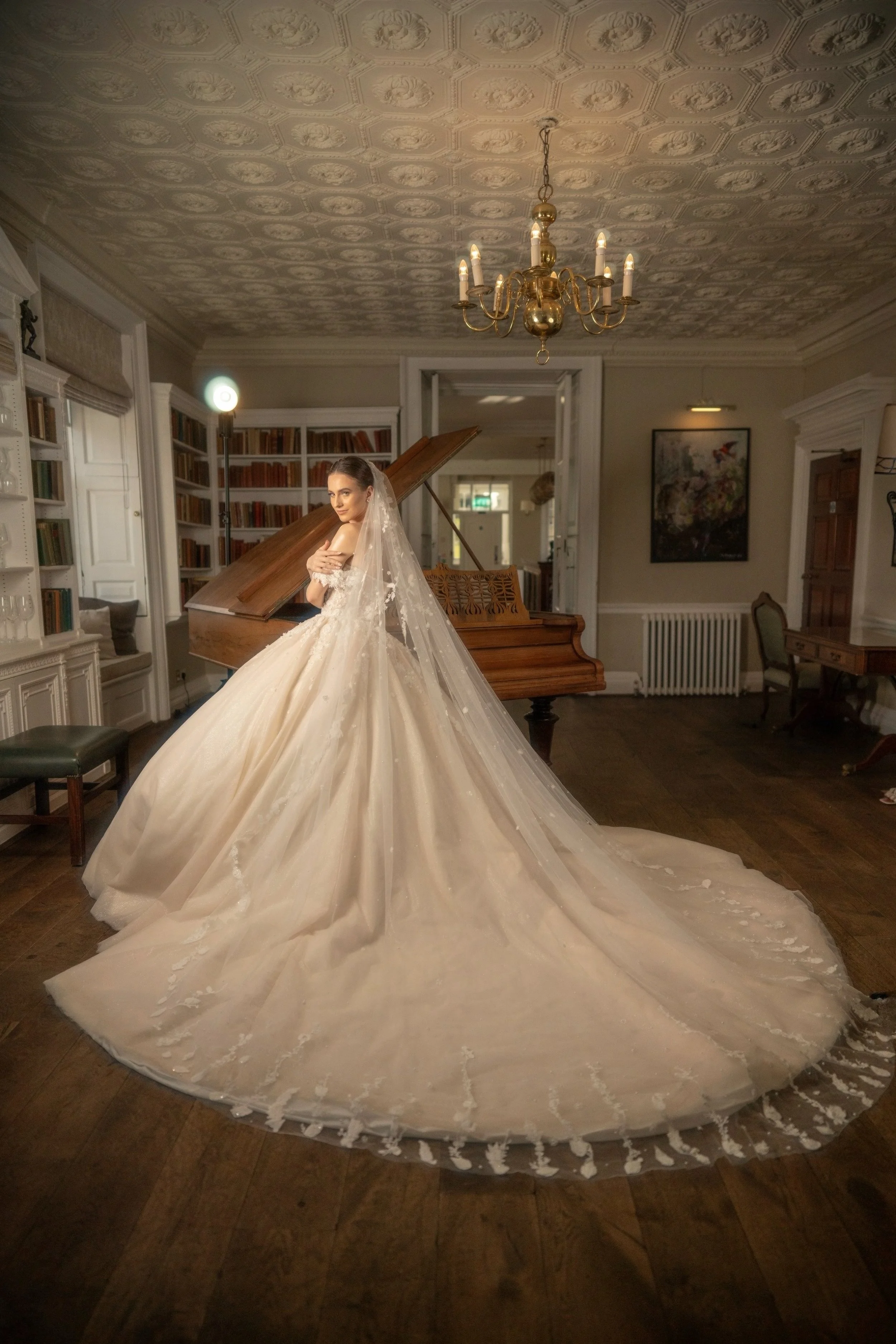 Bride in a wedding gown with a long train standing in a library room near a grand piano.