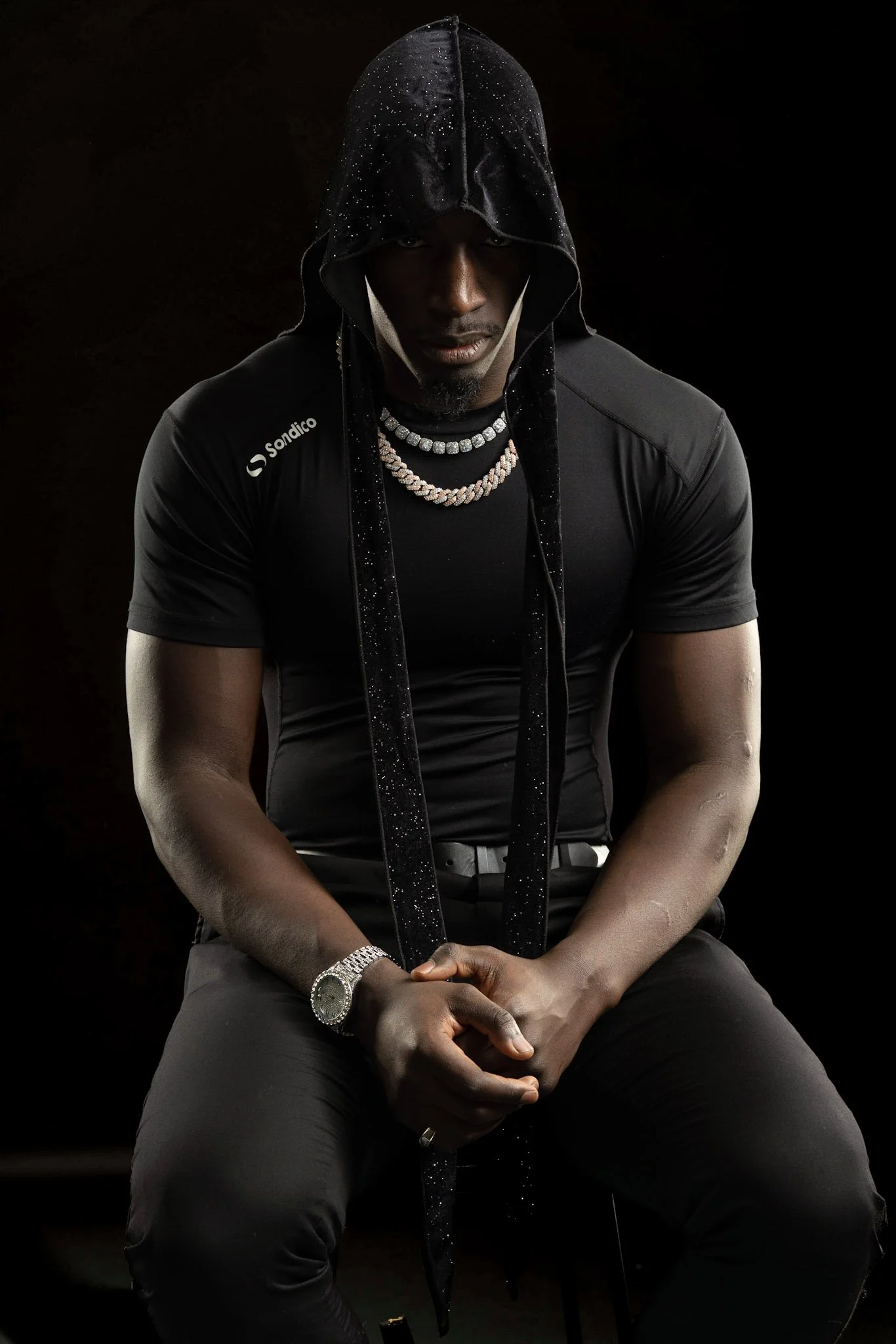 A man sitting against a dark background, wearing a black hoodie with sparkles, multiple necklaces, a watch, and rings, with his hands clasped.
