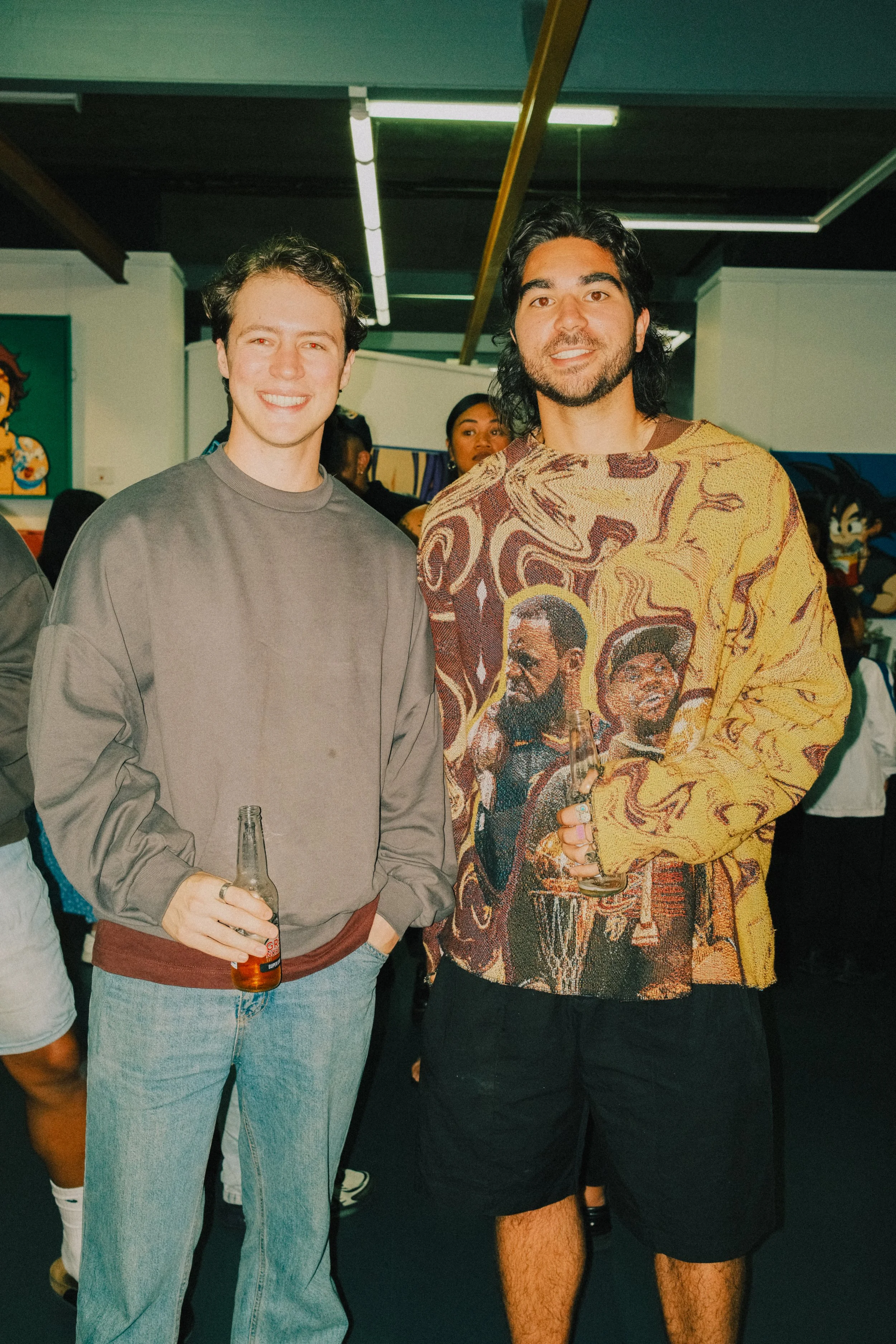 Two men smiling and holding drinks at a social event, with people and colorful artwork in the background.