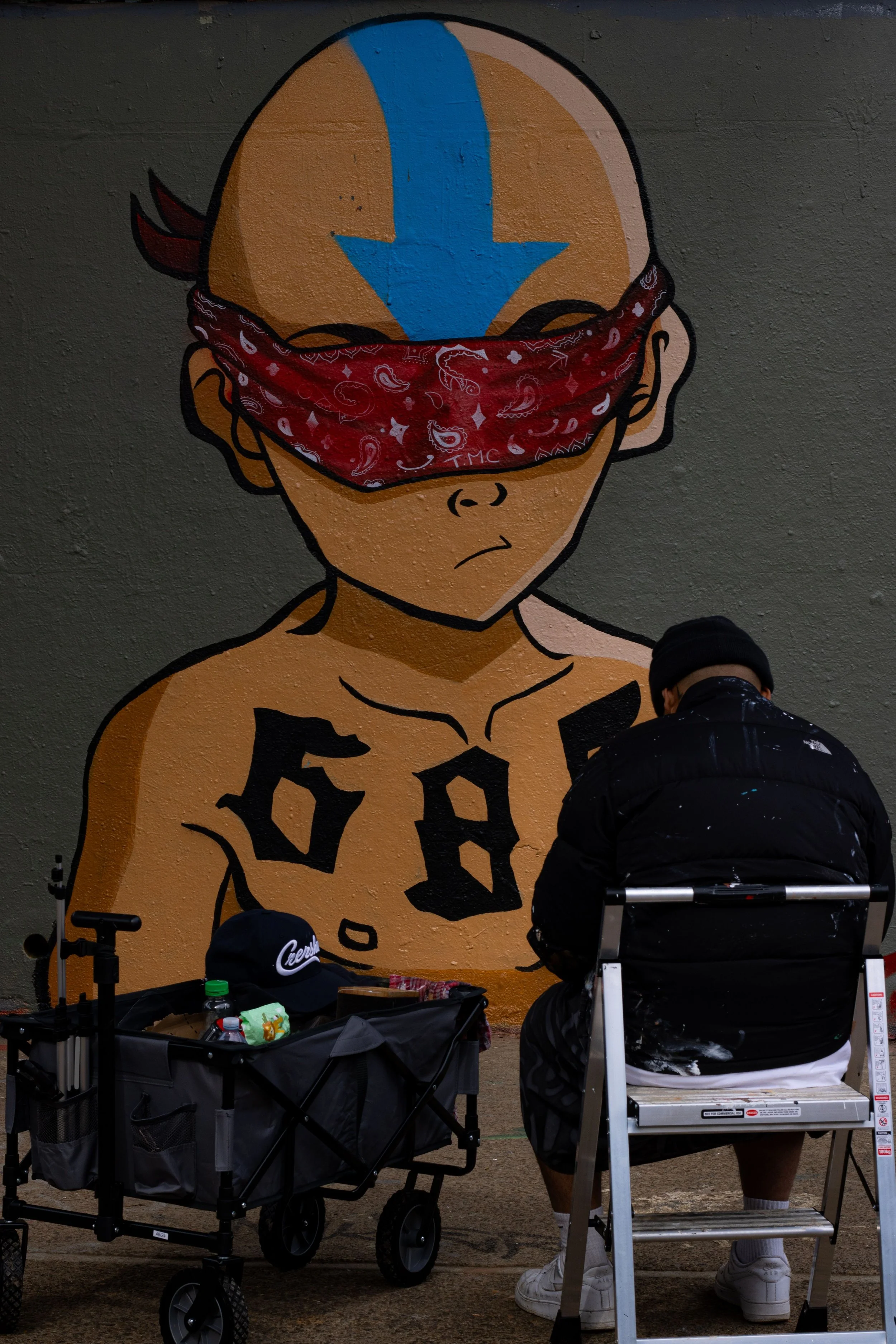 Street artist creating graffiti of a cartoon boy with a red bandana over his eyes and a blue arrow painted on his head, with the word 'BOY' on his chest, on a gray wall.