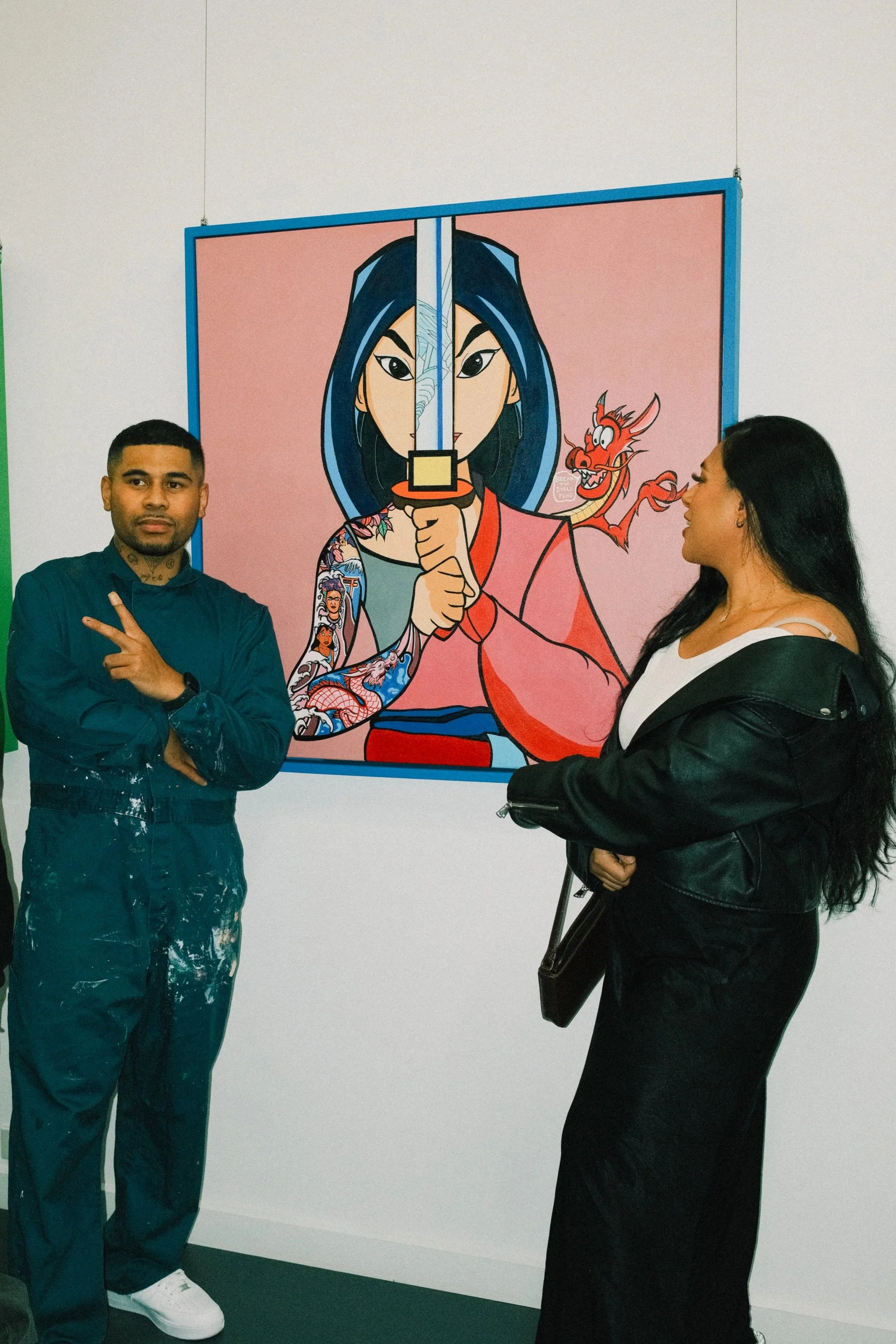Two people standing in front of a colorful artwork depicting a woman with blue hair holding a sword and a small red dragon, with various tattoos on her arms.