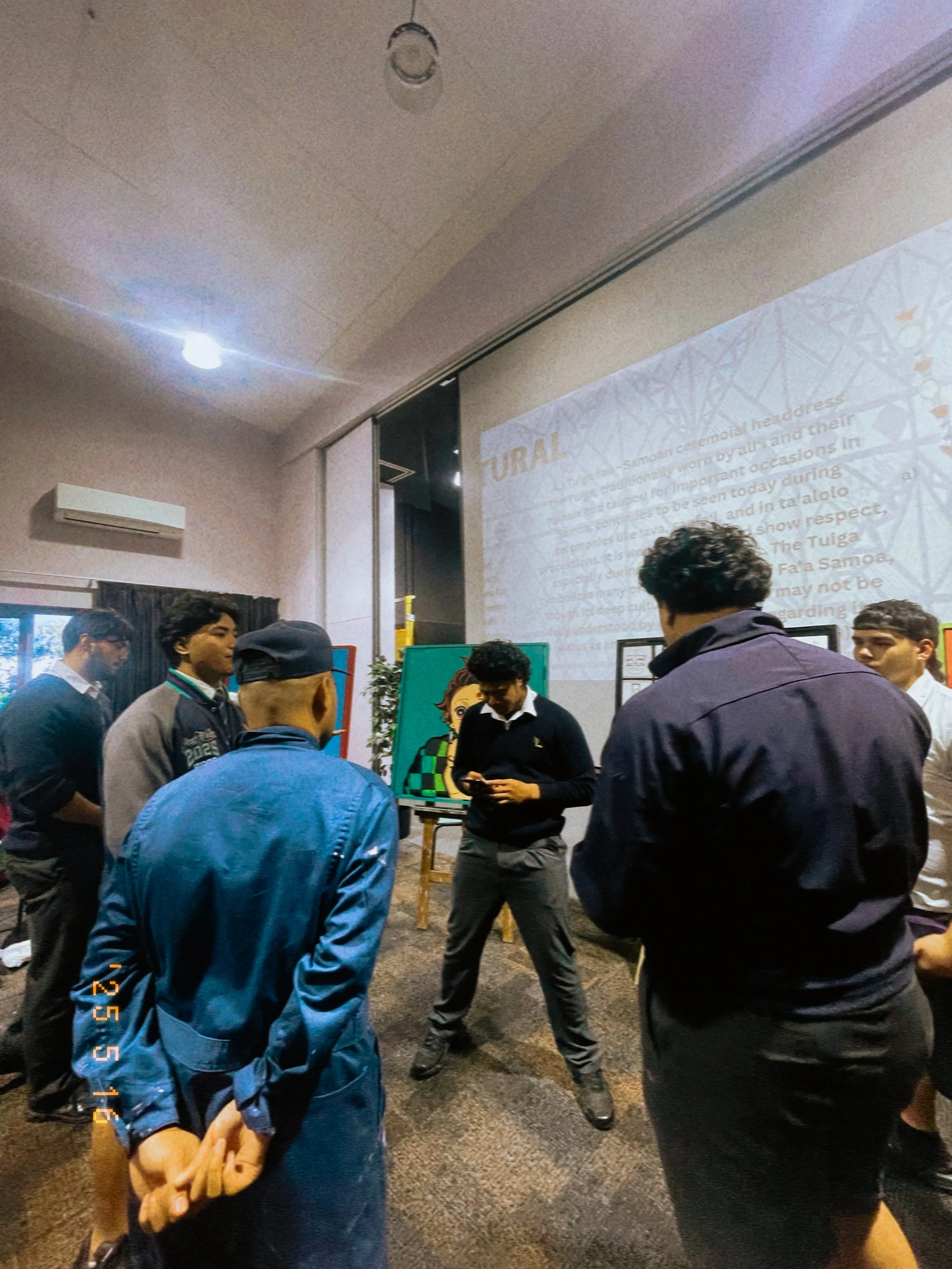 A group of young men are standing and talking in a room with a projection screen displaying information about Fa'a Samoa, the traditional Samoan ceremonial dress, in the background.
