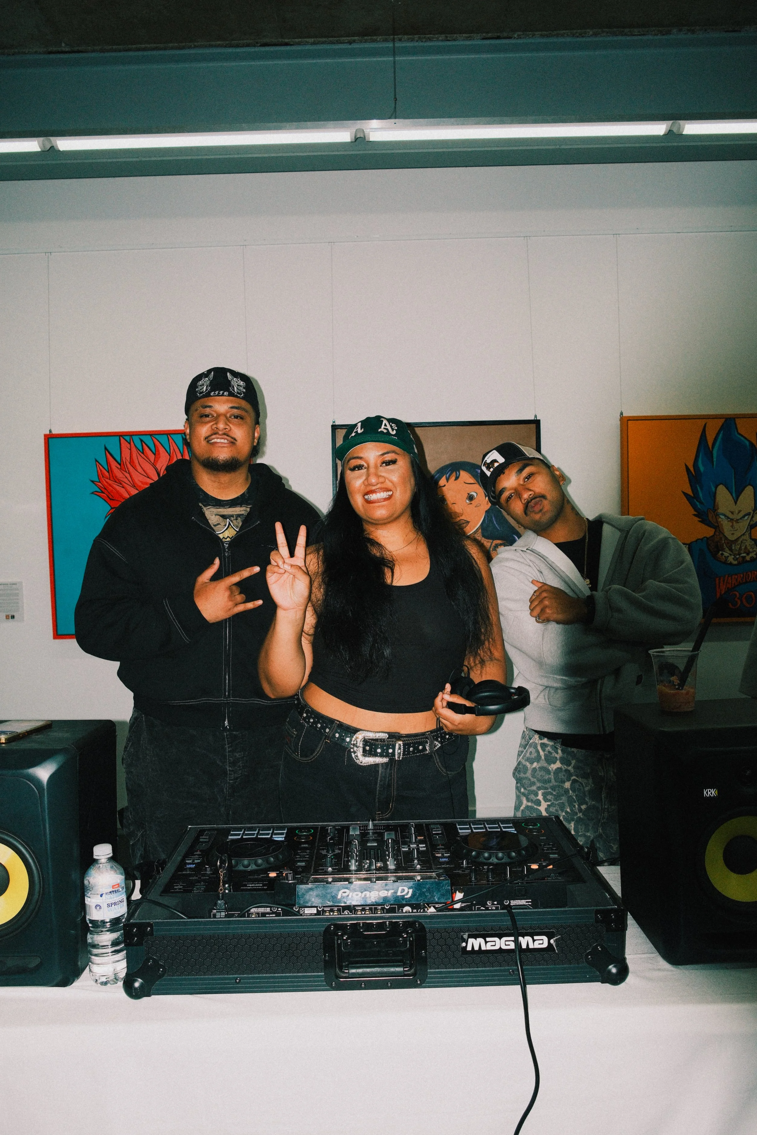 Three people standing behind DJ equipment, smiling, with colorful artwork in the background.