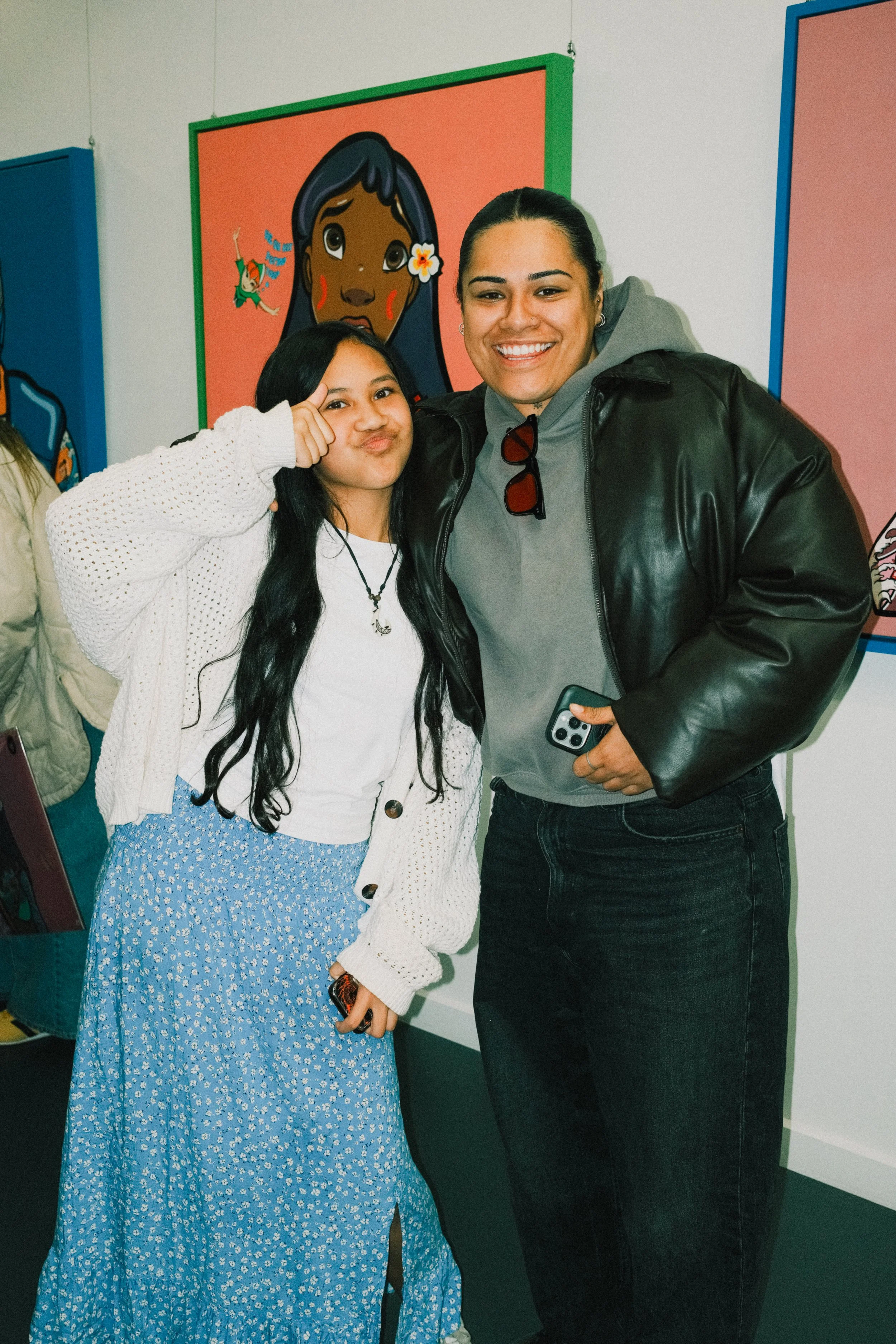 Two people smiling and posing for a photo in front of colorful artwork. The person on the left is a girl with long black hair, wearing a white sweater and a blue skirt. The person on the right is a woman with dark hair tied back, wearing a gray hoodi