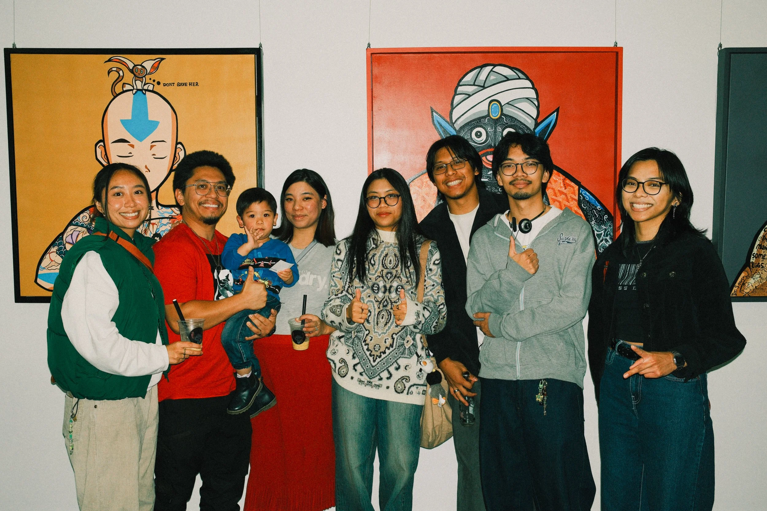 Group of nine people, including a young child, posing in front of colorful artwork at an art gallery. They are smiling, with some giving thumbs up or pointing, and holding drinks.