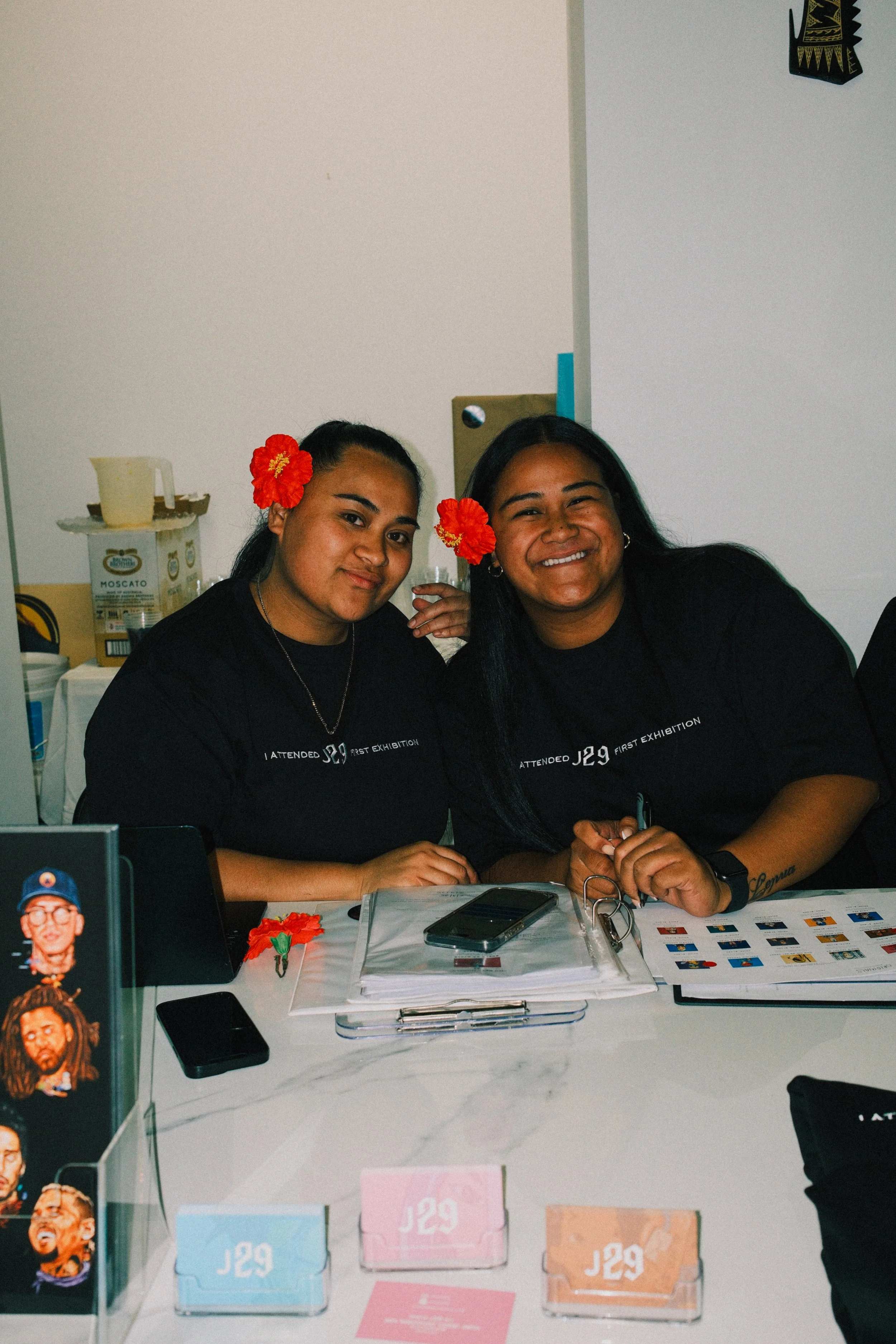 Two women sitting at a table, smiling, wearing black shirts with writing, red flower hair accessories, and surrounded by papers, a phone, and promotional materials in what appears to be an indoor event or exhibition.