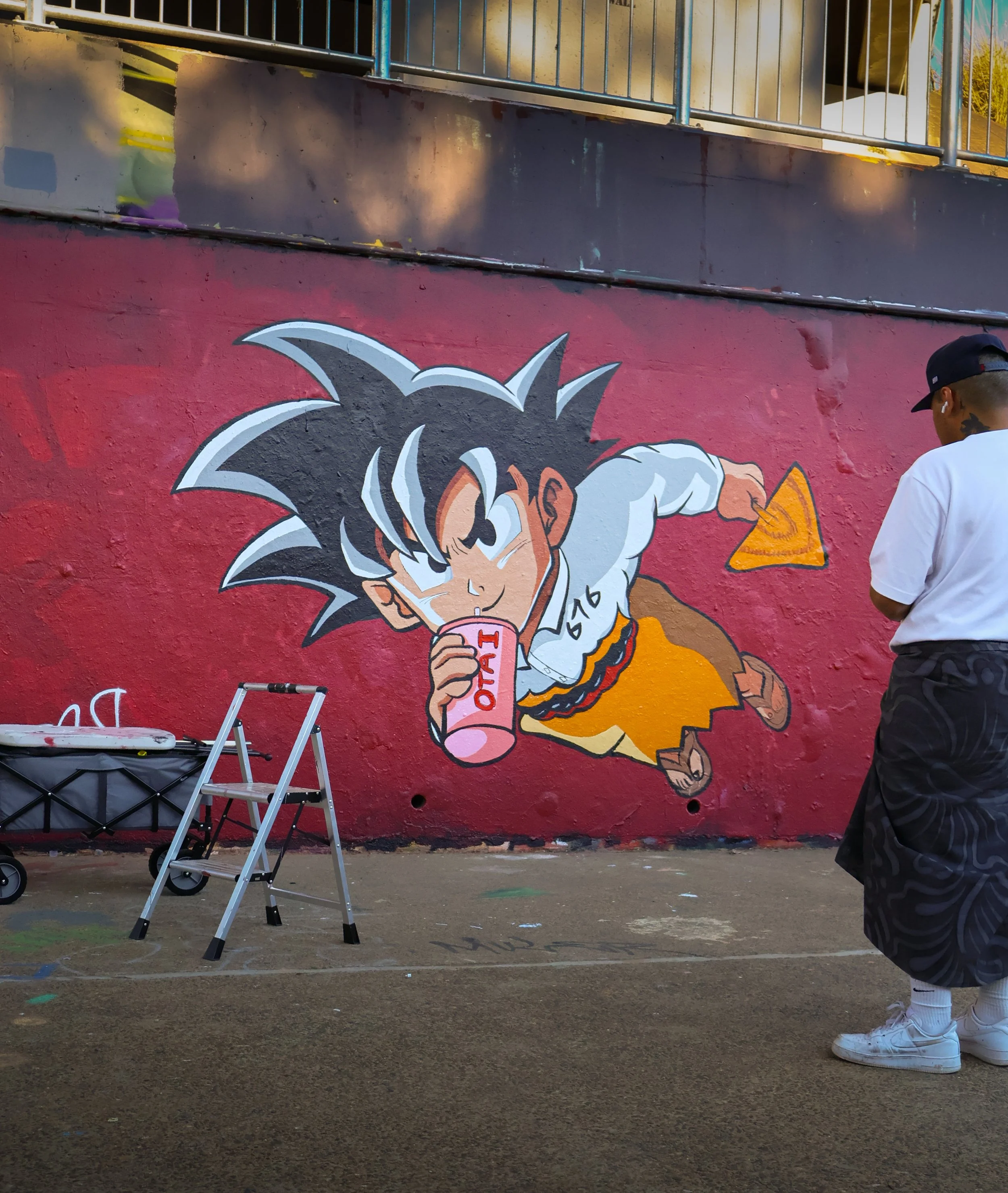 Street art mural of a cartoon character resembling Goku from Dragon Ball Z, dressed in orange and white, drinking from a pink cup labeled 'OAT' on a pink background.