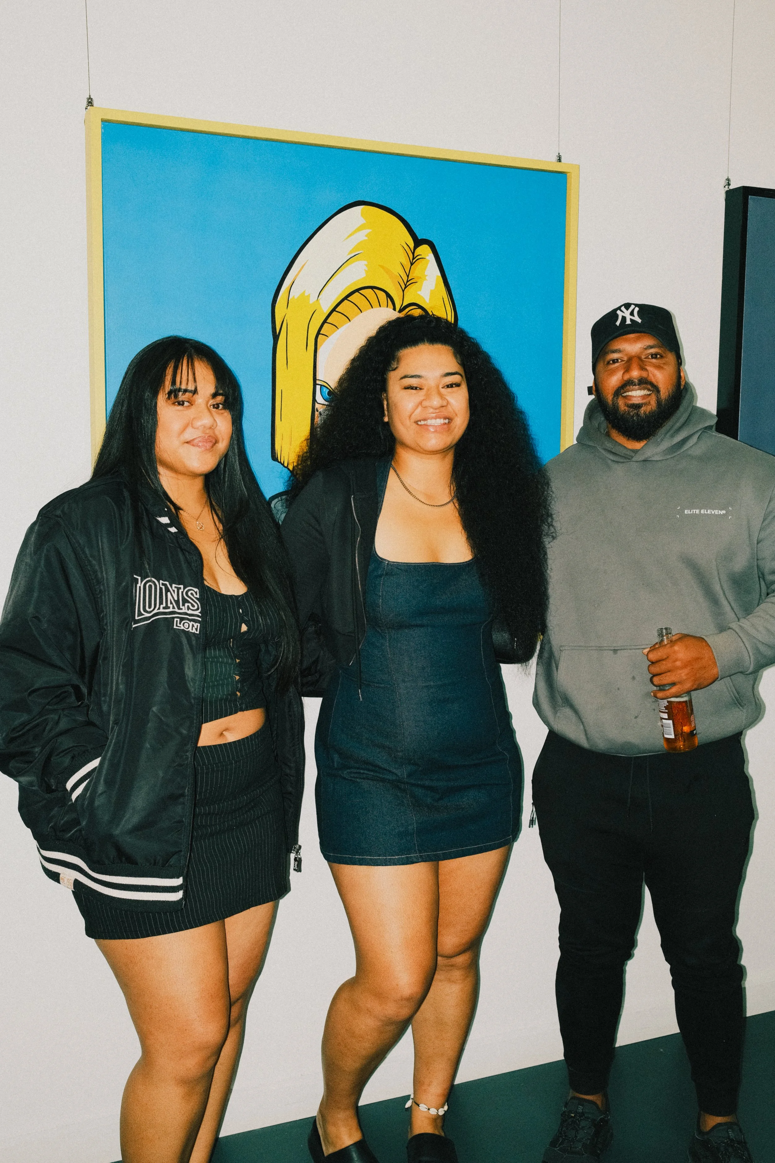 Three people standing in front of a colorful cartoon portrait, smiling at the camera. The woman on the left is wearing a black jacket over a black and striped crop top and black skirt. The woman in the middle is wearing a dark dress and has long, cur