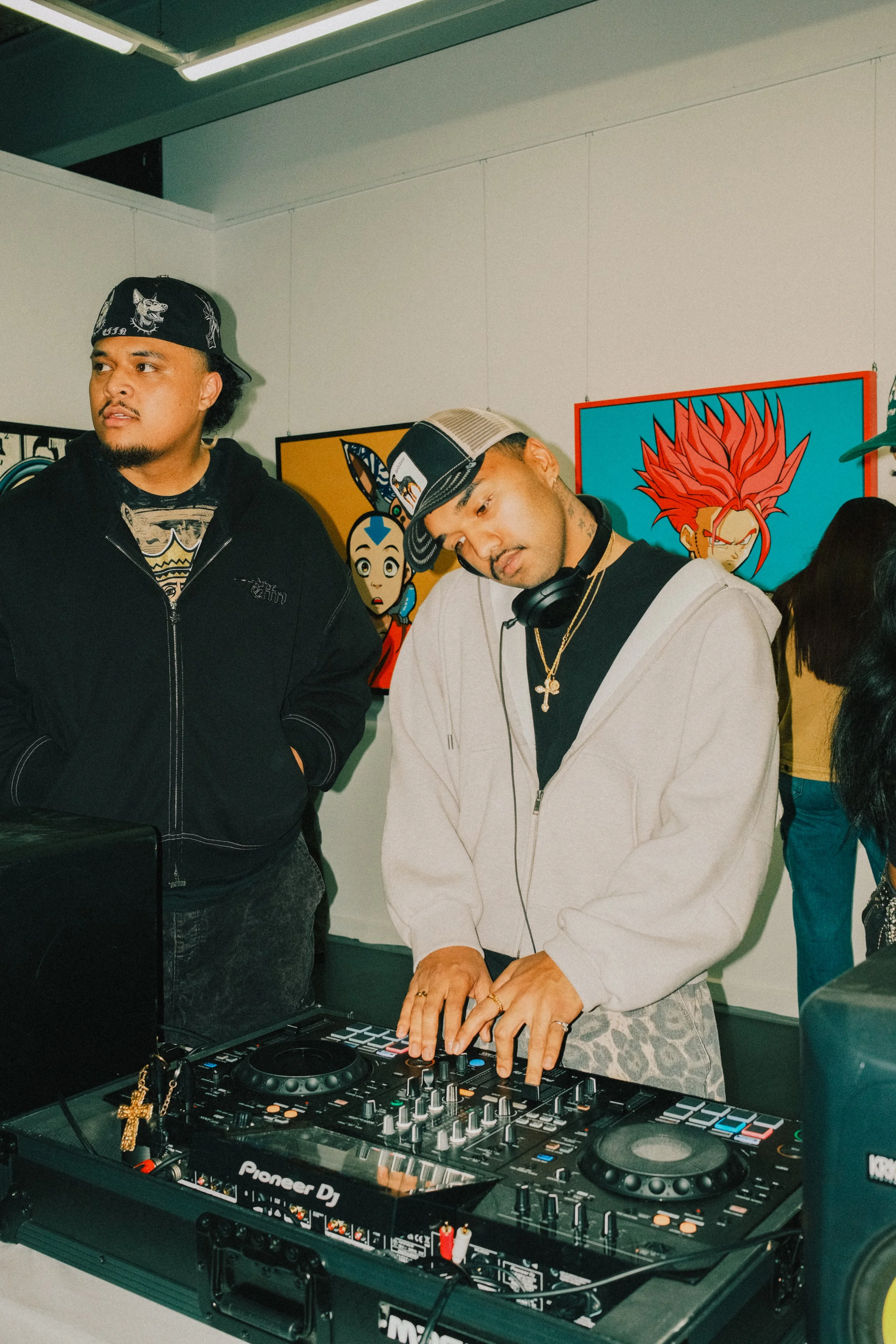 A DJ wearing a beige hoodie, black shirt, and cap with headphones around neck, mixing music at a turntable, with two other men nearby in an art gallery.