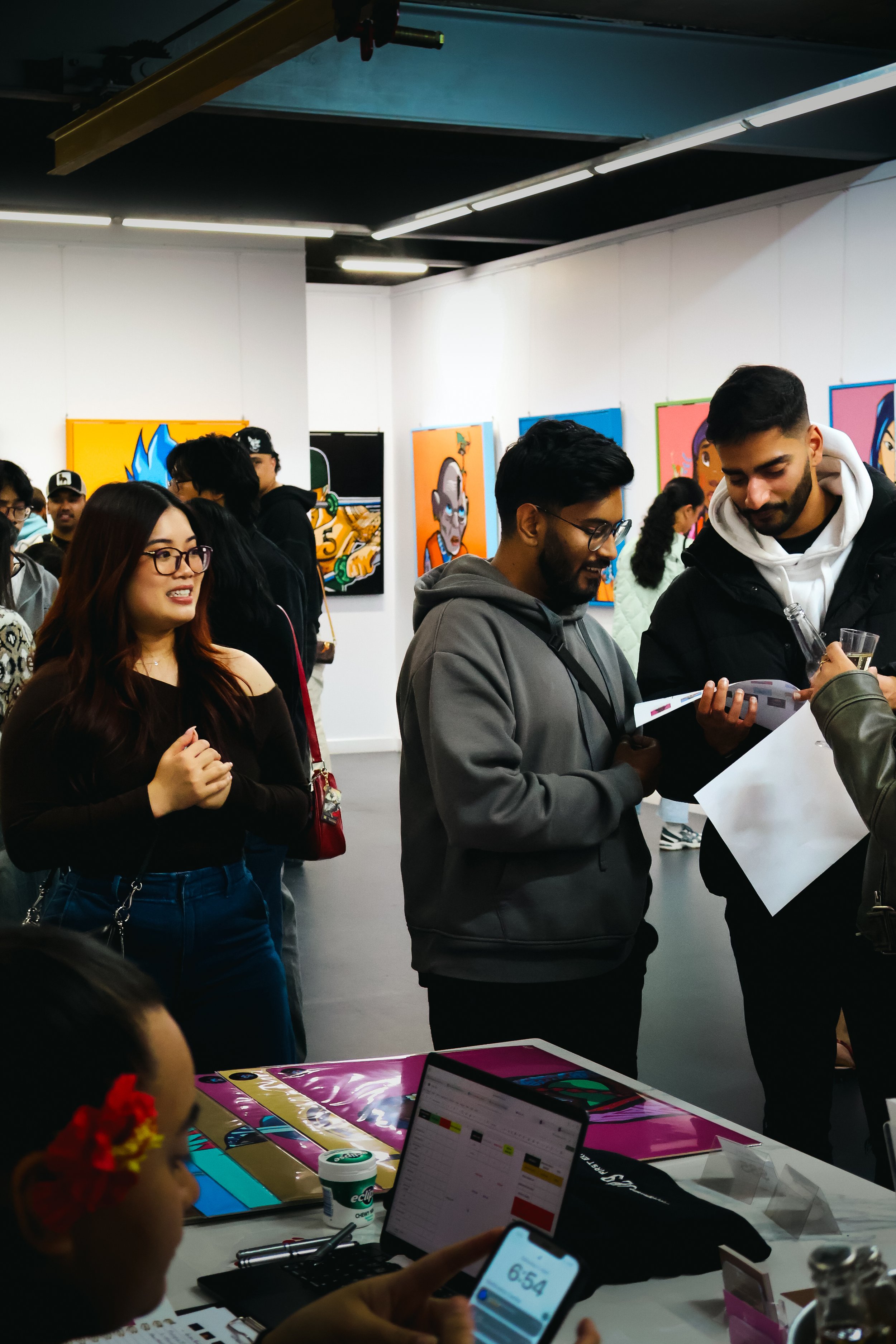 People gathered in an art gallery, viewing colorful artworks on the walls, with some engaged in conversation and others observing the art.