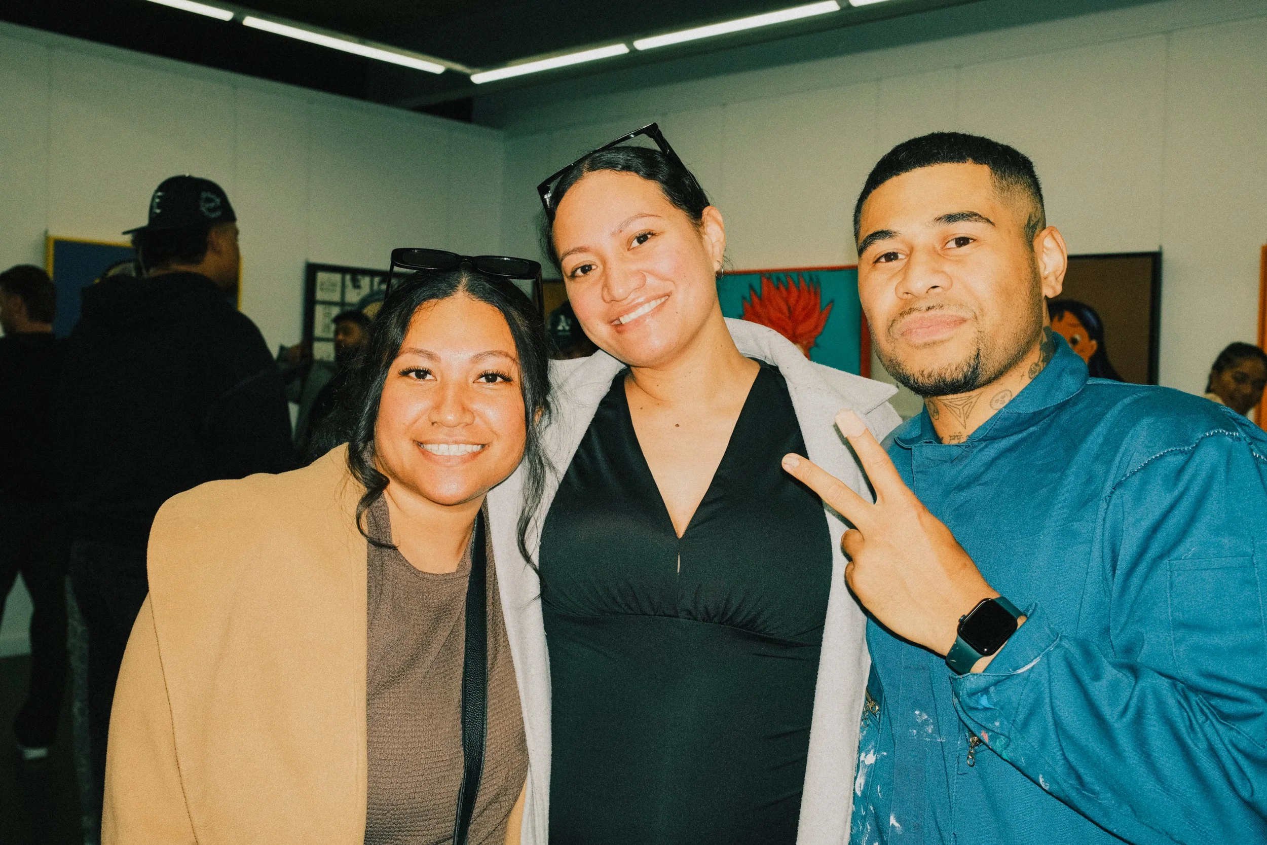 Three young adults smiling and posing together indoors, with artworks on the wall behind them.