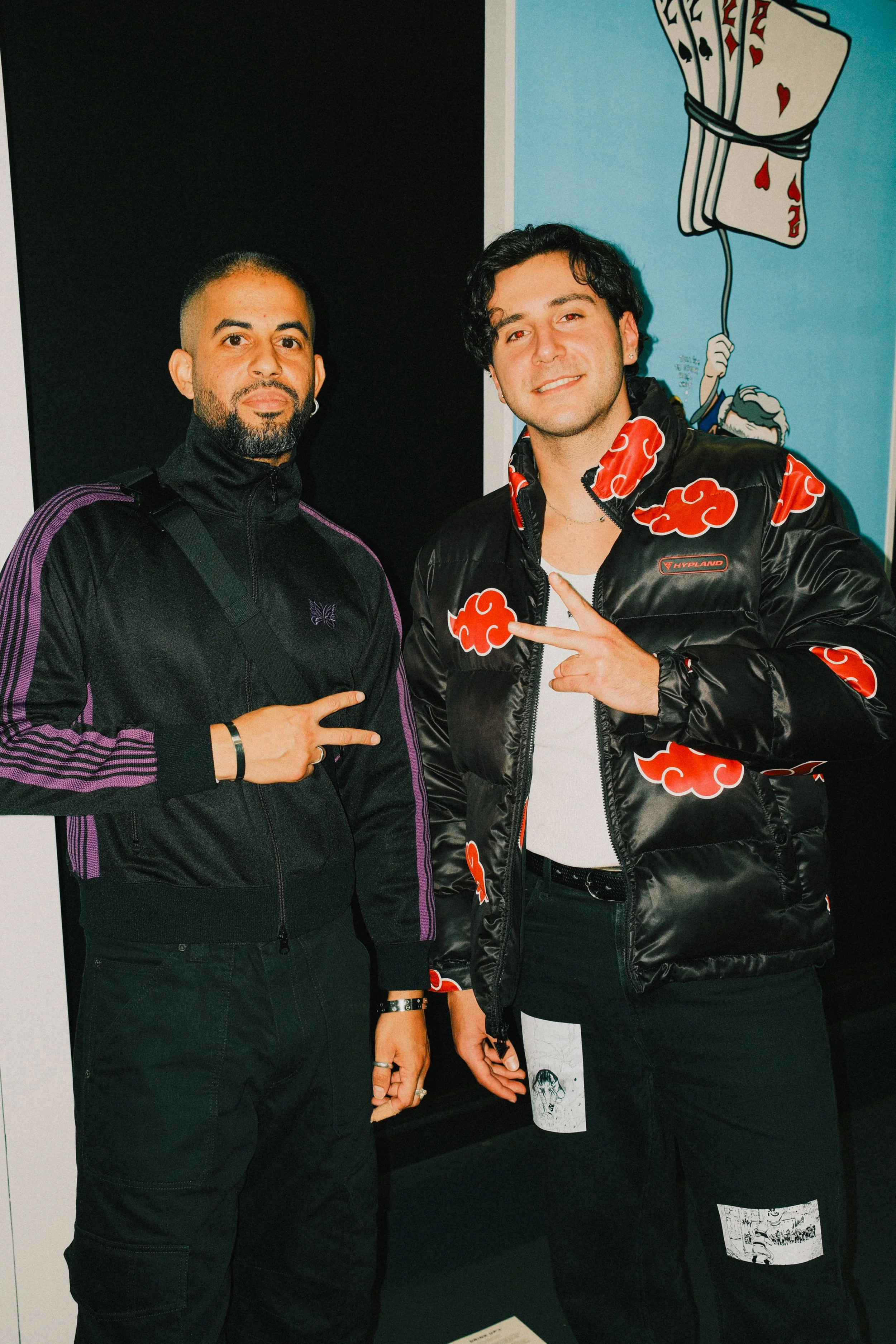 Two men standing indoors, both making peace signs with their fingers. The man on the left has a beard, is wearing a black jacket with purple stripes, and has a dark background. The man on the right has curly hair, is wearing a black jacket with red c
