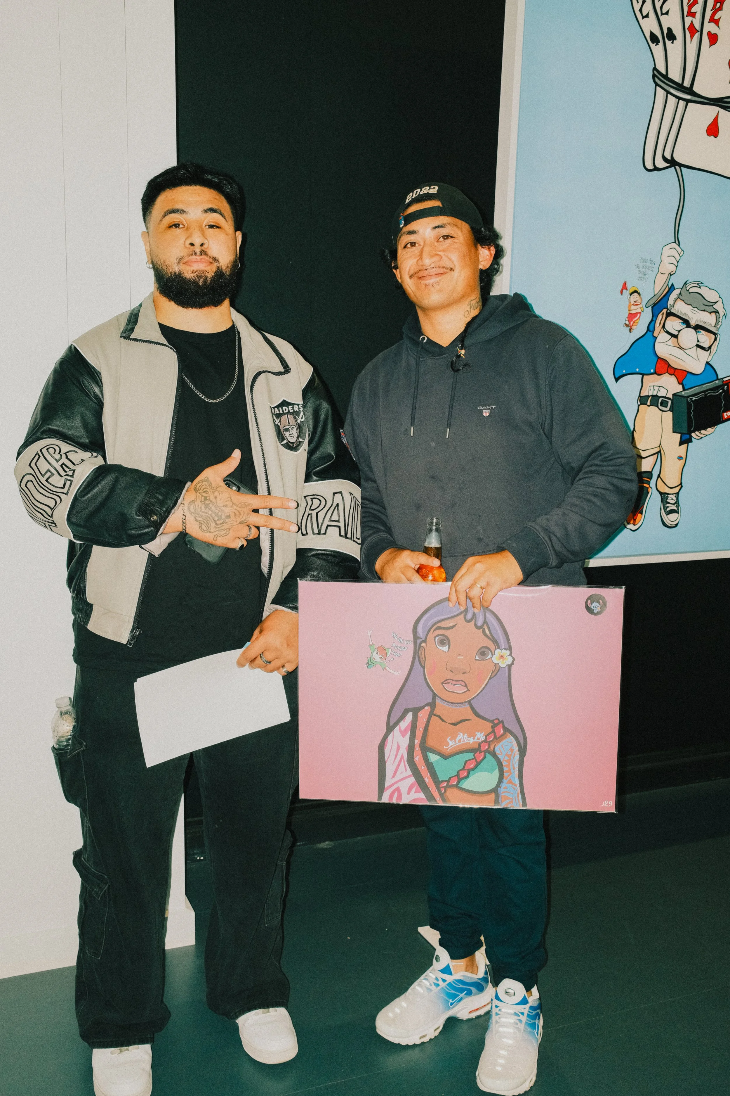 Two men stand together, one holding a bottle and a sign featuring a cartoon girl with purple hair, tattoos, and a flower in her hair, in front of a colorful backdrop with cartoon characters.