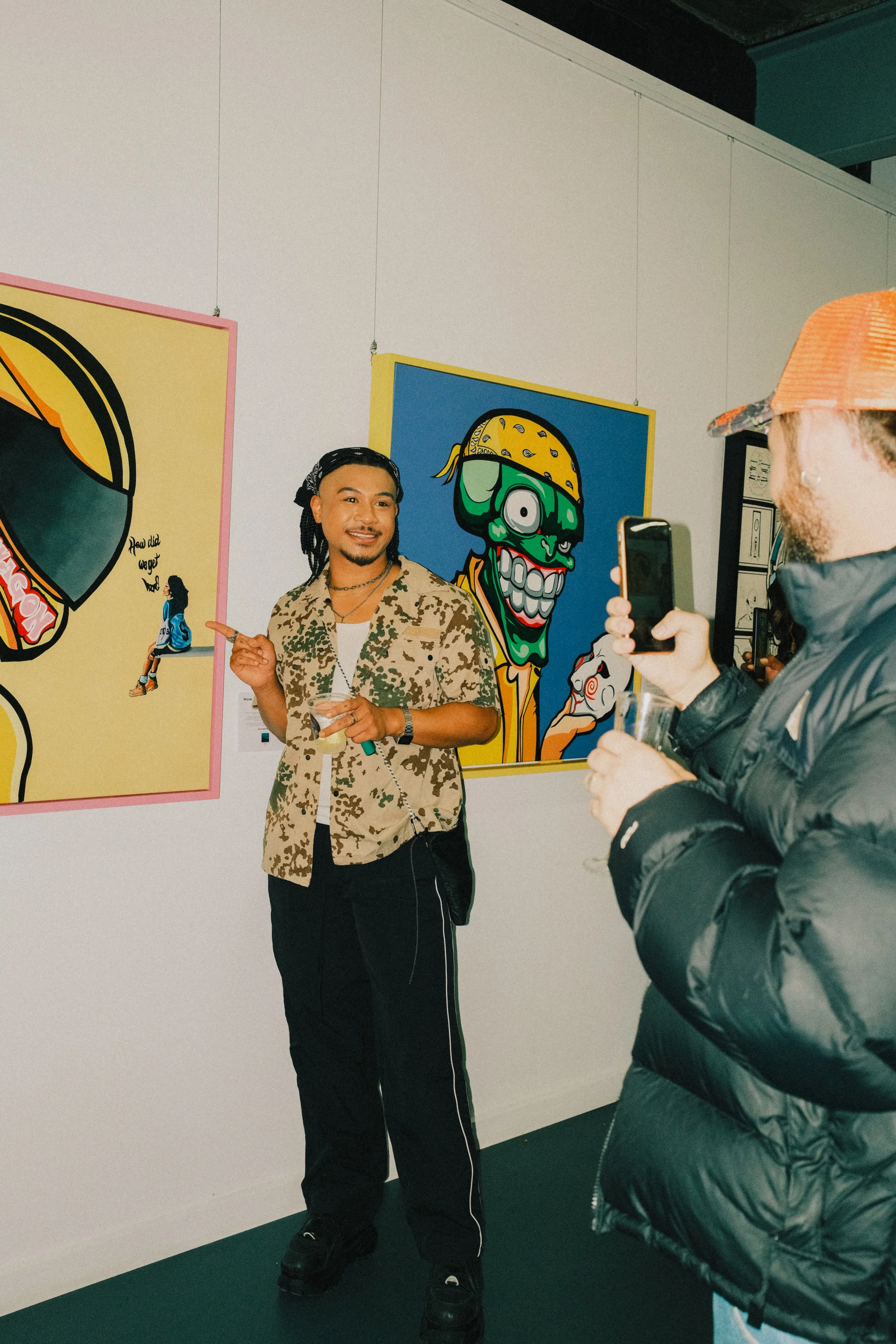 Two people at an art gallery, one person pointing at colorful artwork on the wall, the other person taking a photo or video with a smartphone. The artwork includes a cartoonish face with sunglasses and a bandana and another with a large, smiling mout