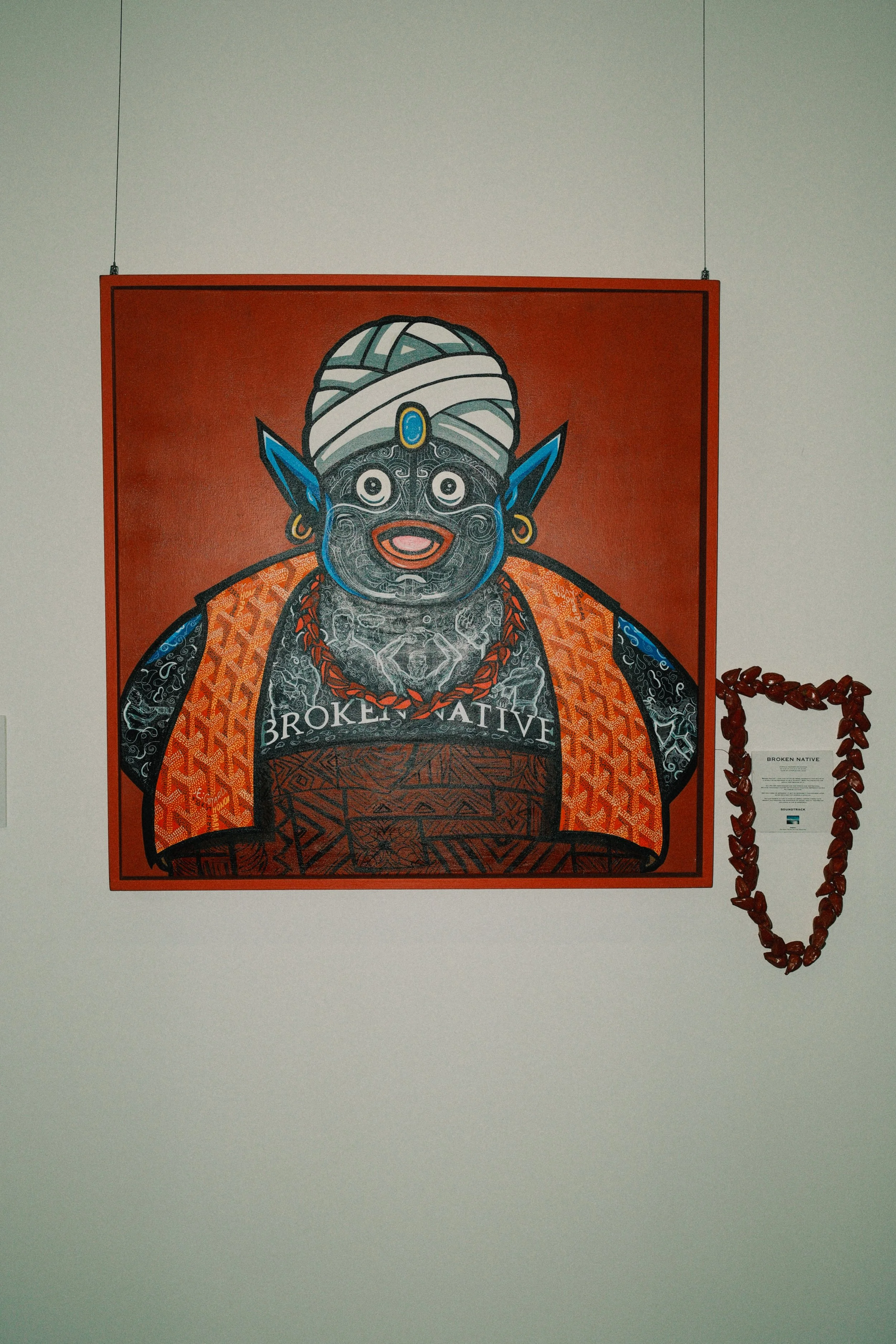 Colorful artwork of a figure with a blue face, big eyes, and pointy ears, wearing traditional clothing and a white turban, with the words 'BROKEN NATIVE' written on it, hung on a white wall with a red necklace hanging beside it.