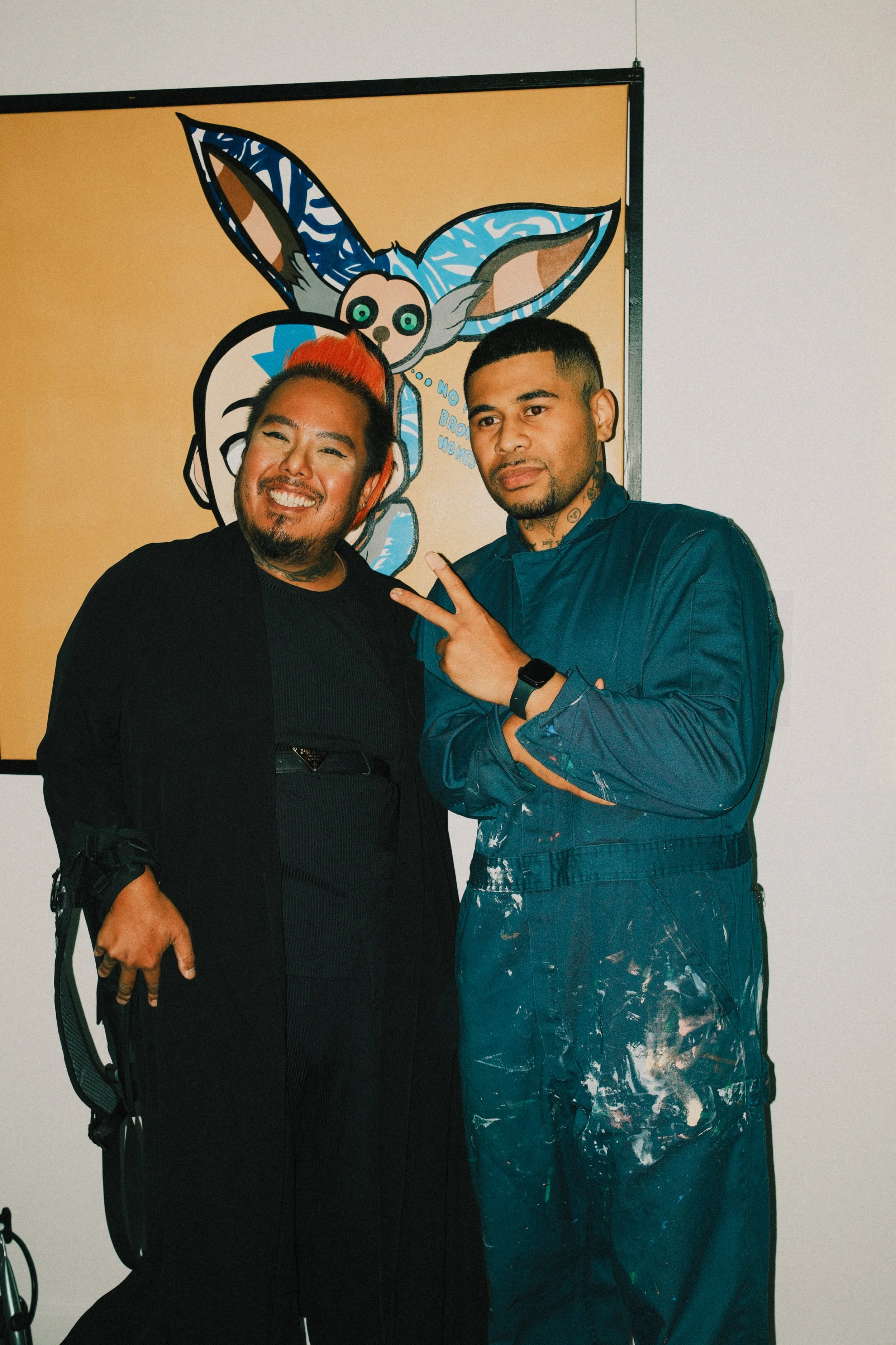 Two men pose together in front of a colorful cartoon artwork featuring a creature with large ears and a skull-like face. One man is smiling with his eyes closed and the other making a peace sign with his fingers.