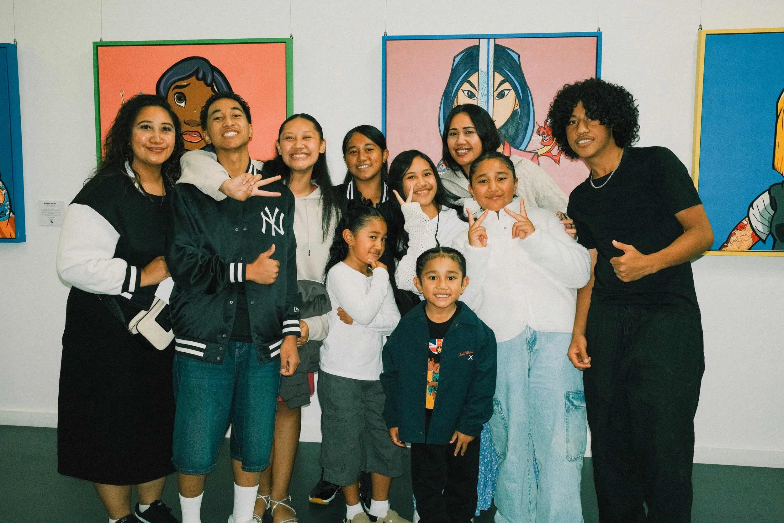 A group of children and adults posing together in front of colorful artwork at an art exhibit, smiling and making peace signs.