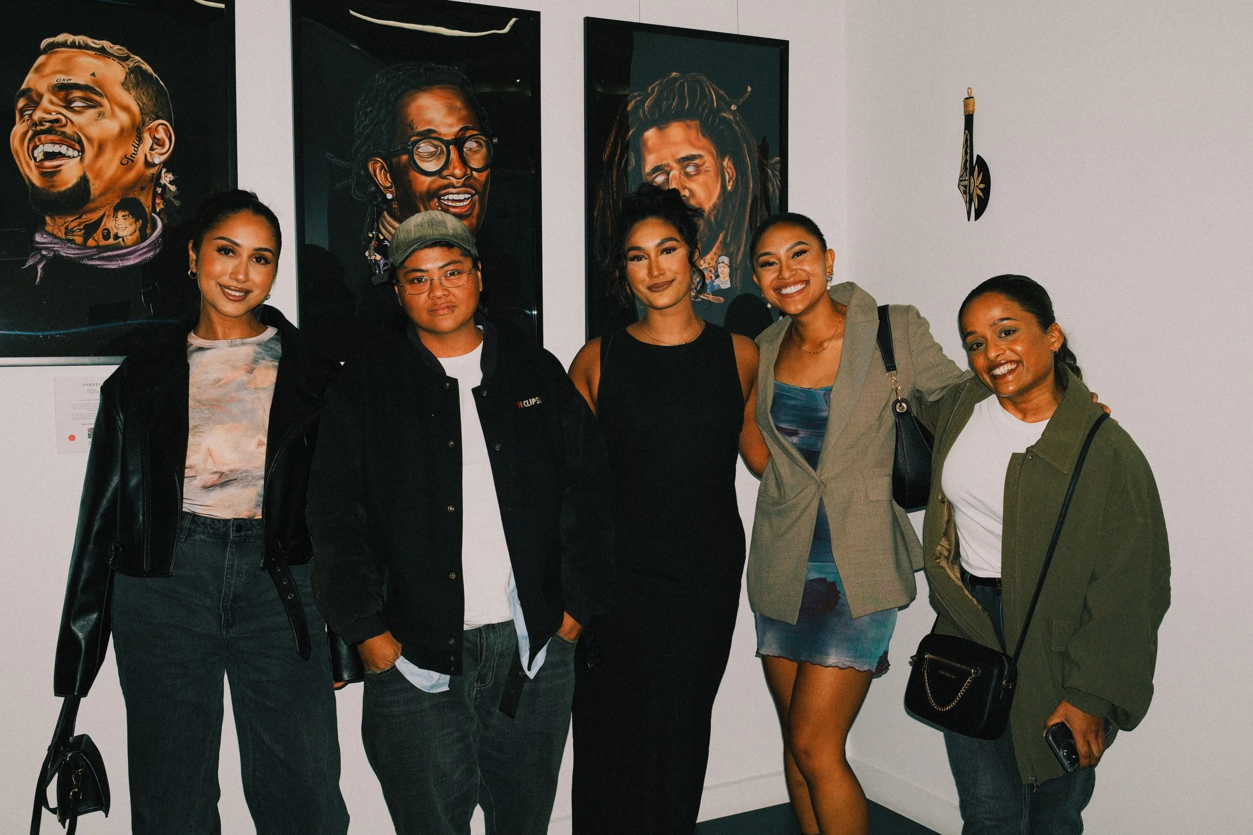 Six women standing together at an art gallery, smiling at the camera with colorful portrait paintings of diverse individuals hung behind them.