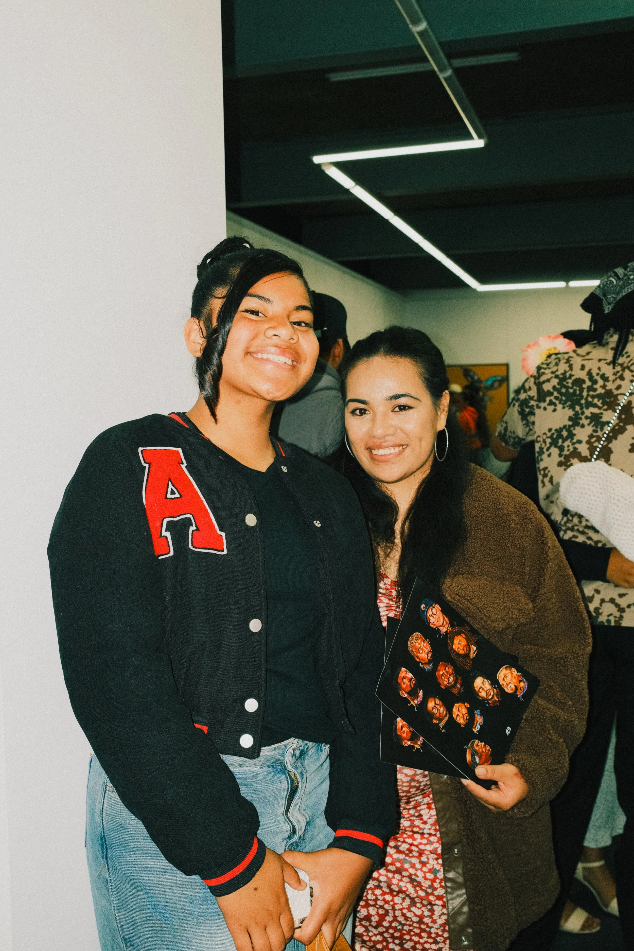 Two women smiling at a social gathering, one holding photo strips with multiple images of herself.