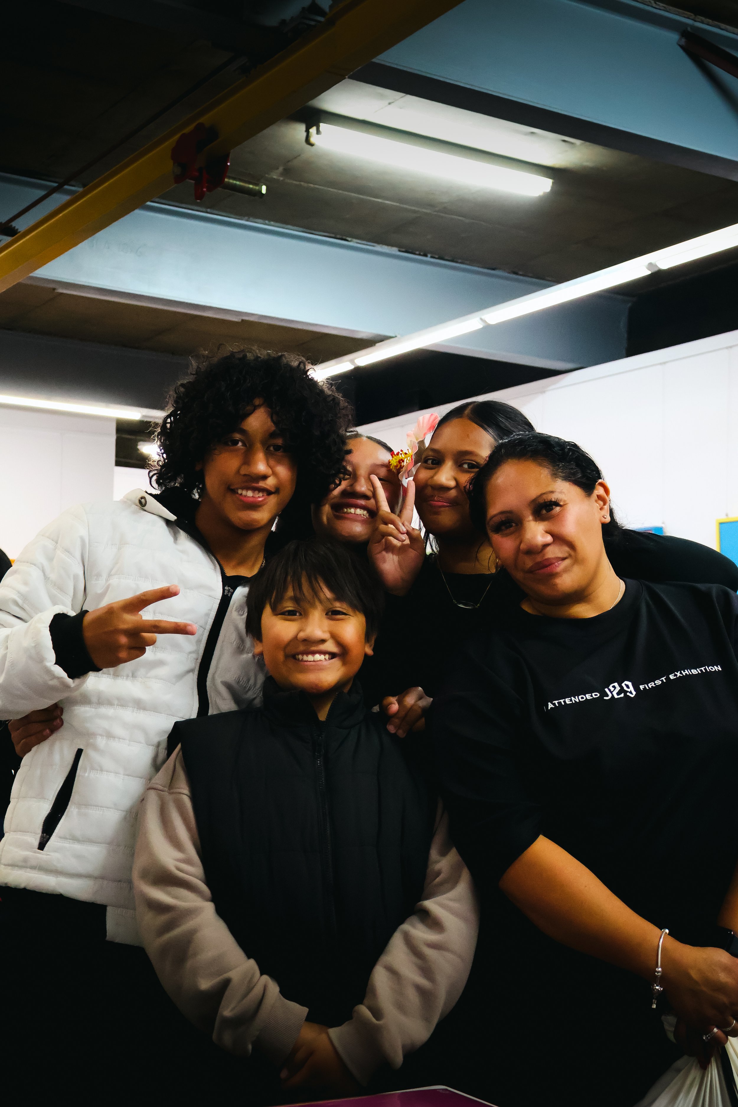 A group of five diverse people smiling and posing together indoors, some making peace signs, with a plain white wall and fluorescent lights in the background.