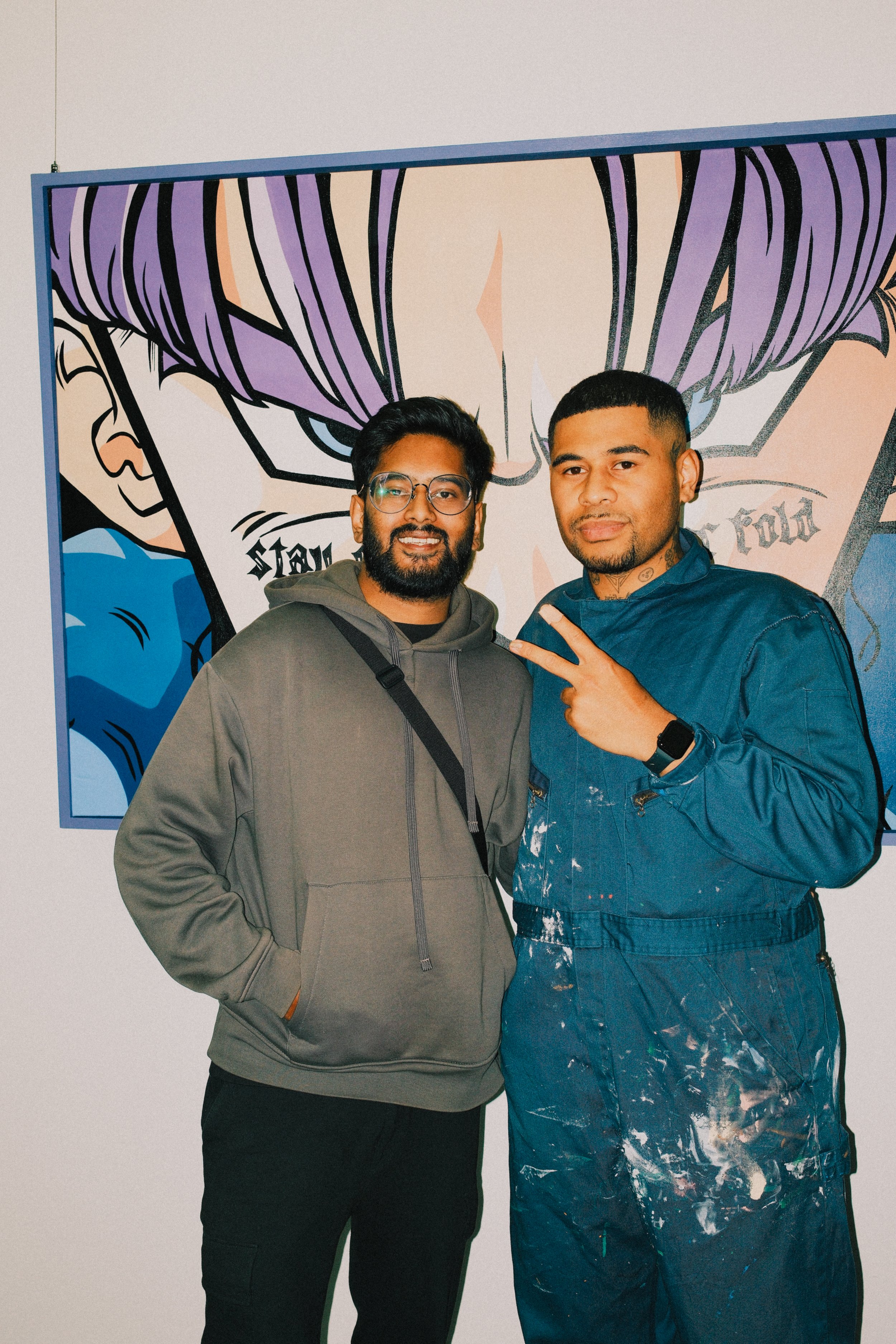 Two men standing in front of a colorful comic-style mural. One is wearing glasses and a gray hoodie, and the other is wearing a blue jumpsuit with paint splatters, making a peace sign.
