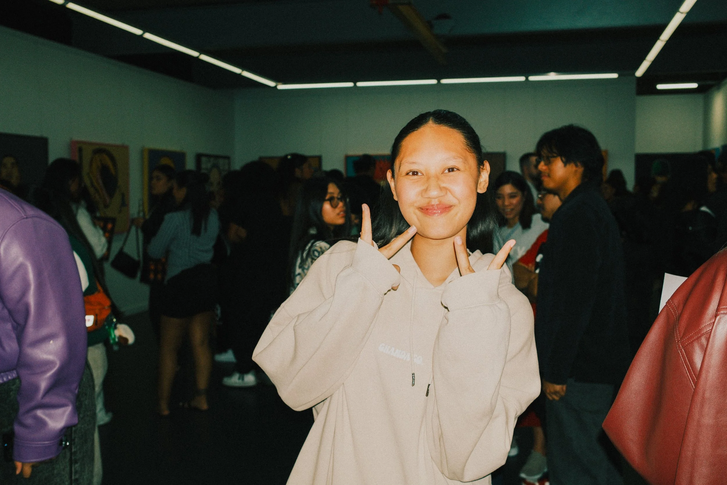 A young girl smiling and making a peace sign with her hands at an art gallery event, with other people and artwork visible in the background.