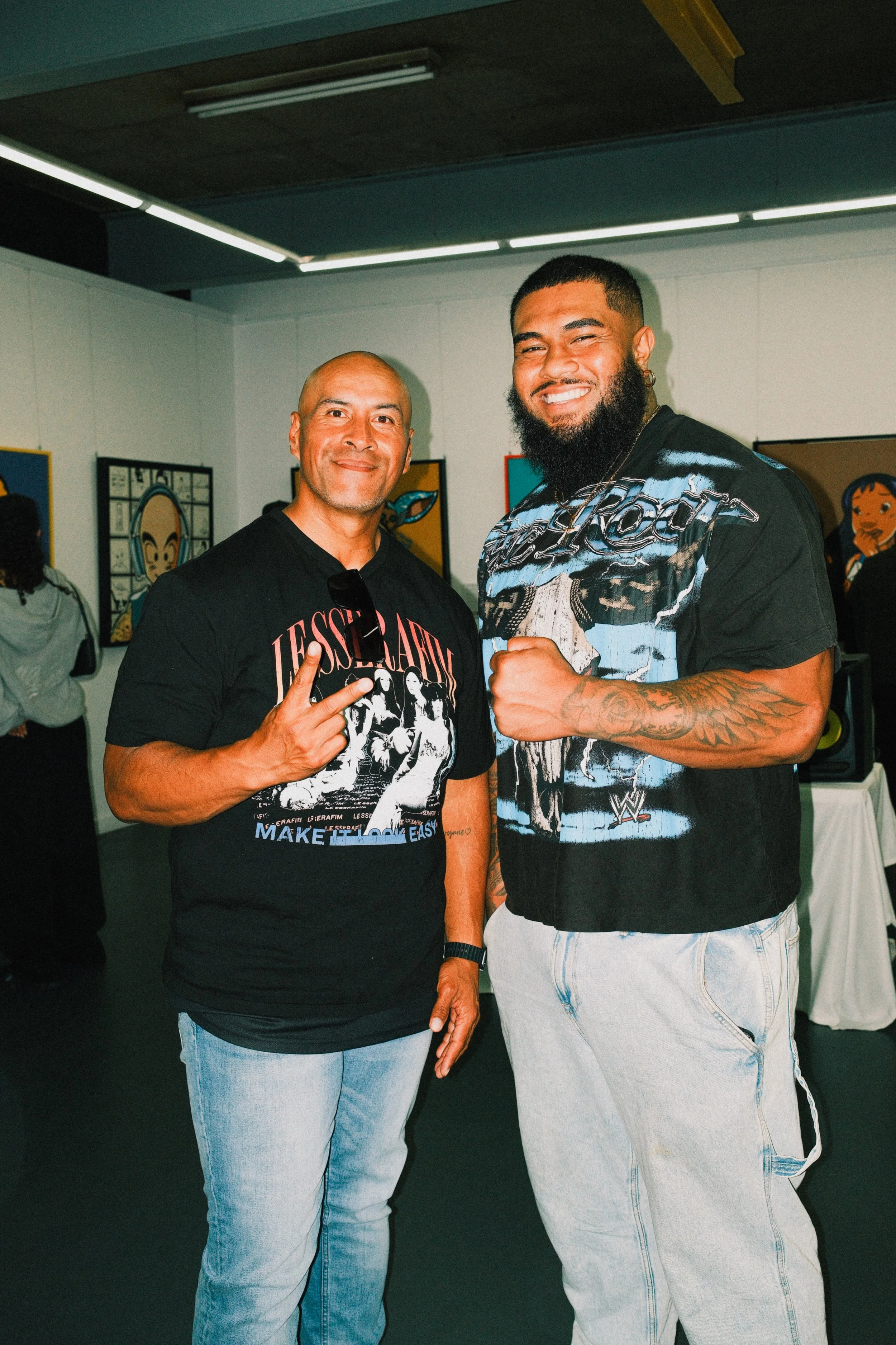 Two men smiling and posing at an art gallery, one making a peace sign and the other with a fist. Artwork is visible on the walls behind them.