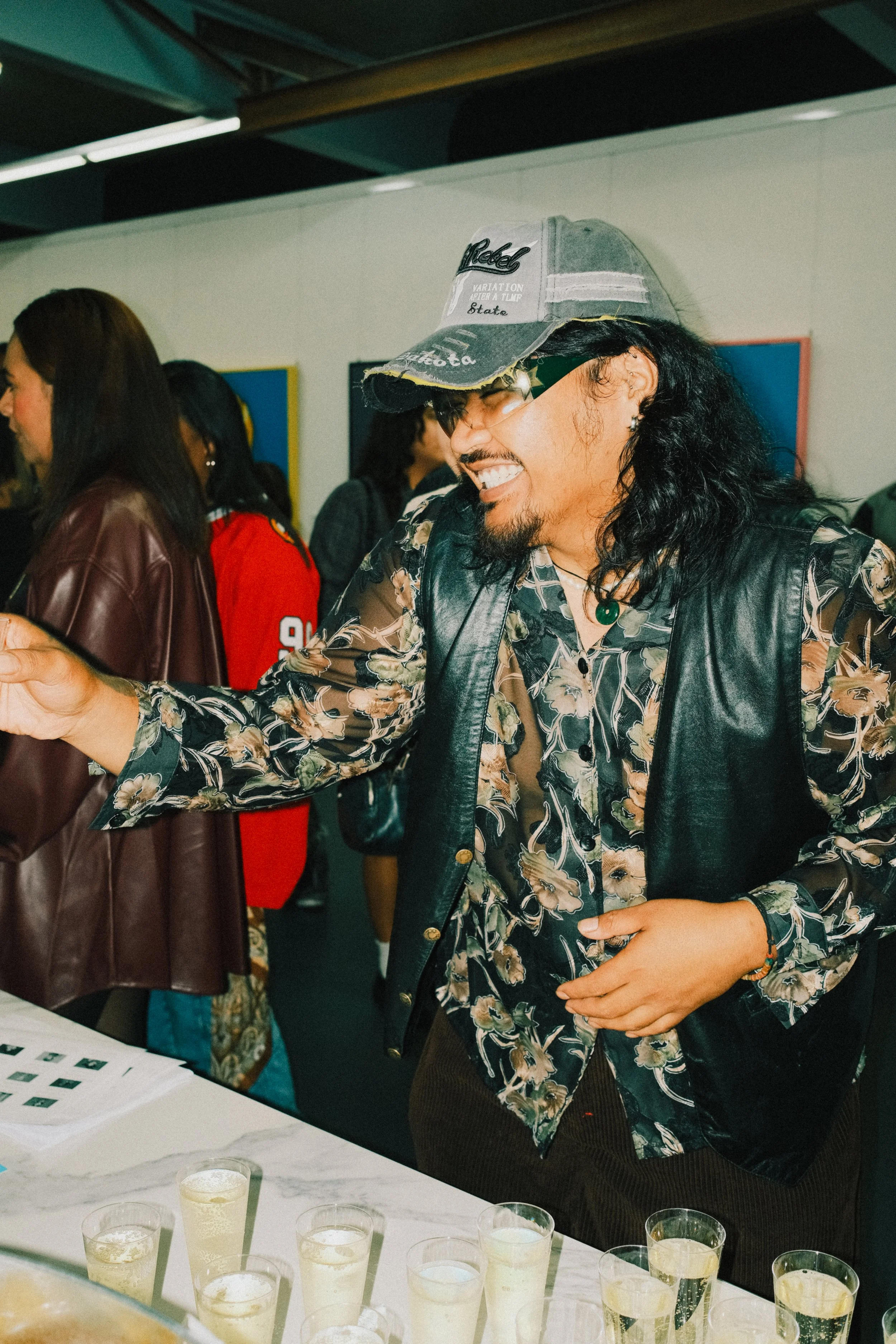 A man with long black hair, a beard, glasses, and a cap is smiling and reaching out towards a table of drinks. He's wearing a patterned shirt and a black leather vest. Several people are in the background at an indoor social event.