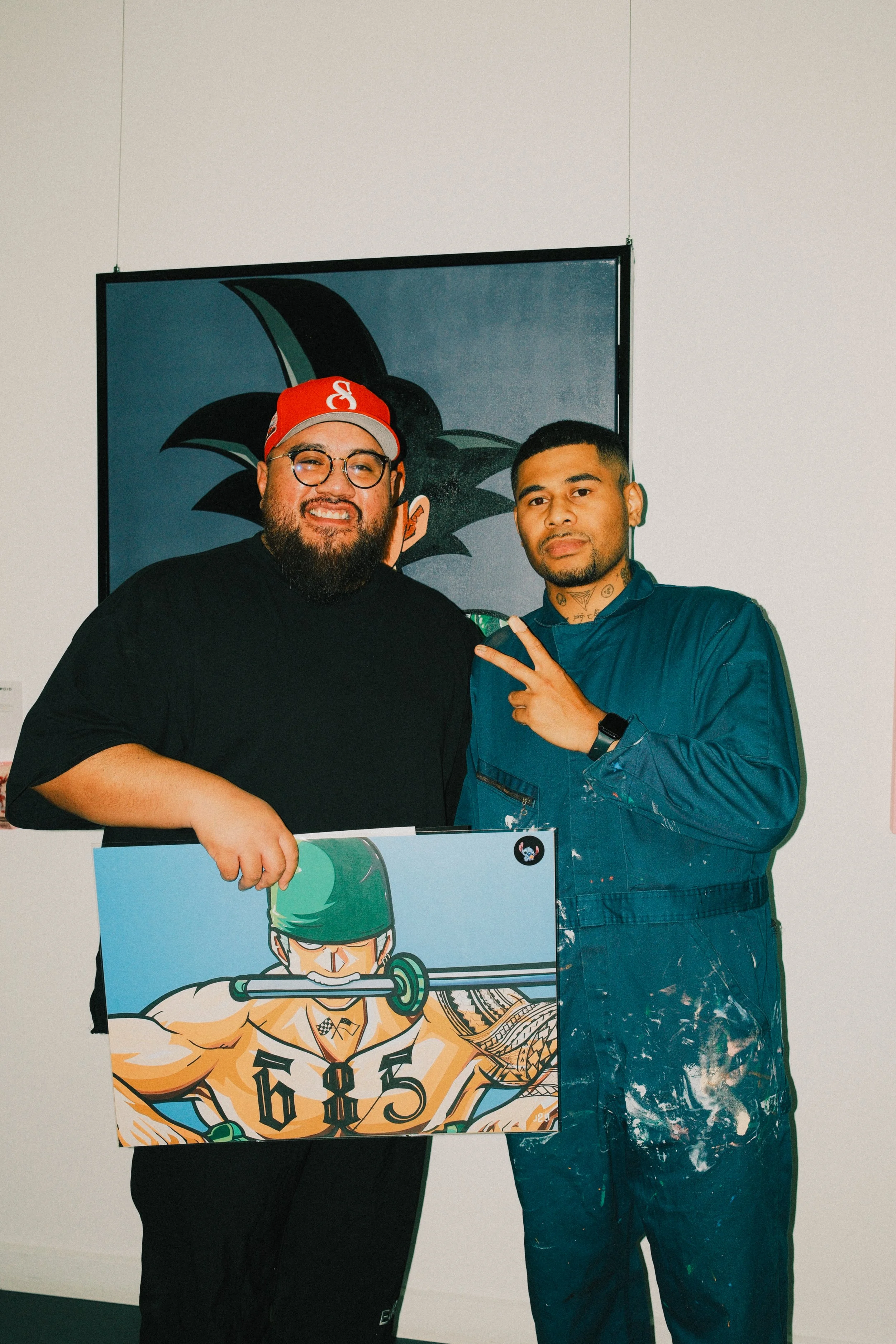 Two men standing in front of a black and blue artwork. The man on the left has a beard, glasses, and is wearing a red cap and black shirt. The man on the right has tattoos on his neck and is wearing a blue jacket with paint stains. They are holding a
