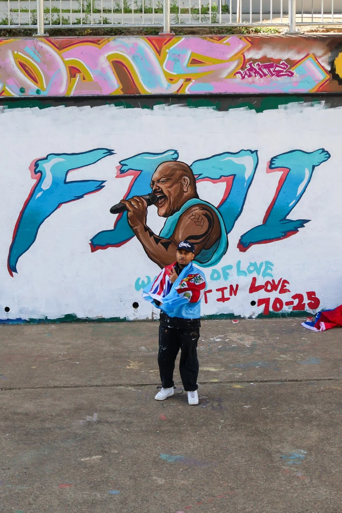 Person standing in front of a large mural of Tupac Shakur singing into a microphone, with colorful graffiti art in the background.